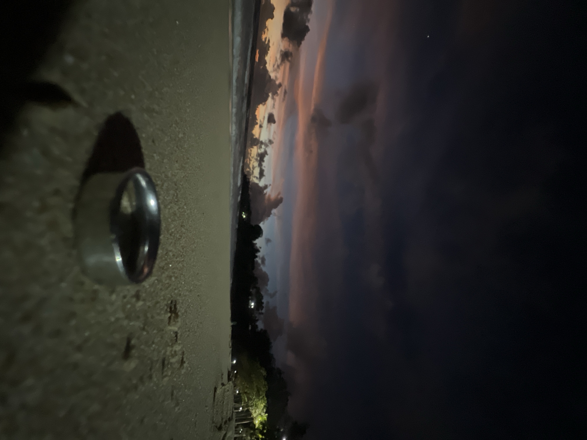 This media file depicts a beach scene at dusk or dawn, with the image rotated 90 degrees clockwise. In the foreground, a silver ring rests on a sandy surface. The sand appears textured and gritty. Beyond the sand, the ocean stretches to the horizon, with gentle waves lapping the shore. The sky is a dramatic canvas of color, with swirling clouds illuminated by the setting or rising sun. Hues of orange, pink, and purple blend with darker shades of blue and grey, suggesting a transition between day and night. The presence of some distant lights, possibly from a town or resort, can be seen along the treeline bordering the beach. The overall mood is serene and contemplative, capturing the beauty of a tropical twilight. The location is identified as Choeng Thale, Thailand.