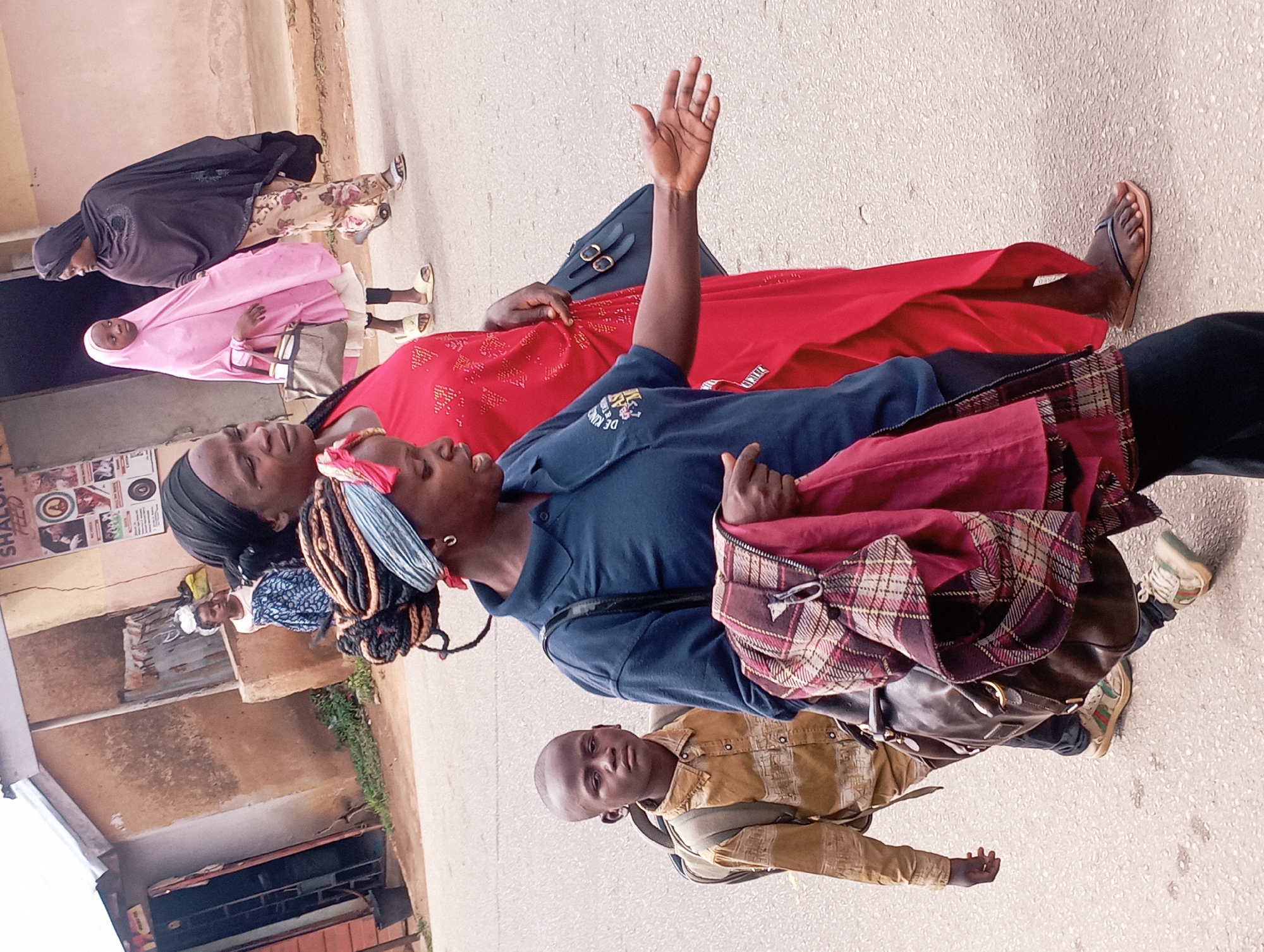 In Jos, Nigeria, a group of people are walking on a paved street. The foreground features a woman in a blue shirt and a patterned headscarf, carrying a plaid blanket and a dark leather bag. To her left, a young boy in a tan shirt and backpack walks ahead, looking directly at the camera with a neutral expression. Behind the woman in blue, another woman in a vibrant red dress walks beside a young girl in a pink hijab and a light-colored dress, carrying a tote bag. Further in the background, a woman in a dark hijab walks with her arm linked with the girl in pink. The buildings in the background have a rustic, earth-toned facade, suggesting a developing urban environment. The overall scene captures a moment of daily life on a street in Jos.
