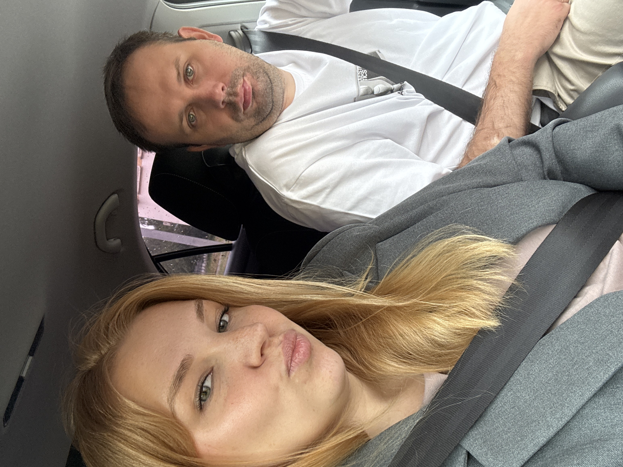 The image is a selfie taken from inside a car. A woman with blonde hair and a grey blazer is in the foreground, making a duck face. Her seatbelt is visible. To her right, a man with short brown hair and a white t-shirt is visible, also wearing a seatbelt. The interior of the car, including the headrest and a portion of the ceiling, is visible in the background. Through the window, trees and buildings can be seen, suggesting an outdoor environment within a populated area. The image quality is moderate, with some graininess. The exact location in the city of Zhukovo, Russia, cannot be determined from the visual information alone.