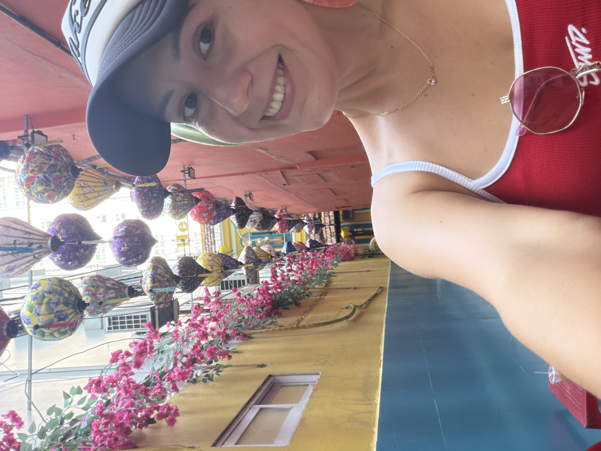 A woman with a tan complexion and dark hair is taking a selfie in a narrow alleyway. She is wearing a red tank top with white trim, a gold necklace with a small pink pendant, and a dark baseball cap. Sunglasses with pink lenses are hooked onto the front of her tank top. Her facial expression is a wide smile, and she is looking directly at the camera.
The alleyway is decorated with numerous colorful, decorative lanterns hanging overhead. The walls of the alley are painted in bright yellow and blue on one side, and pink on the other. Vibrant pink flowers cascade down from the yellow wall. A window with a white frame is visible on the yellow wall. In the background, the alley recedes, showing more buildings and more lanterns. A red table with a drink in a plastic cup is partially visible in the lower left corner of the image. The scene appears to be in an urban environment, specifically identified as Nha Trang Ward, Vietnam.