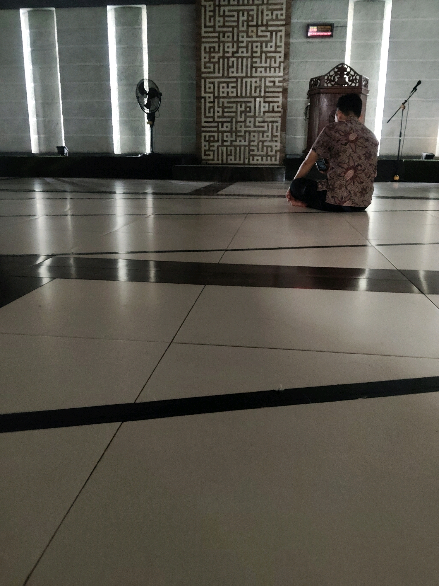 The image shows a man sitting on the floor of a mosque in Mekarmukti, Indonesia. The man is facing away from the camera and is wearing a patterned shirt and dark pants. He is sitting with his legs crossed, and his hands are resting on his lap. The floor of the mosque is tiled with a pattern of light and dark tiles. In the background, there is a large wall with Arabic calligraphy, a wooden podium, a microphone on a stand, and a fan. Vertical light fixtures illuminate the space, casting long shadows on the floor.