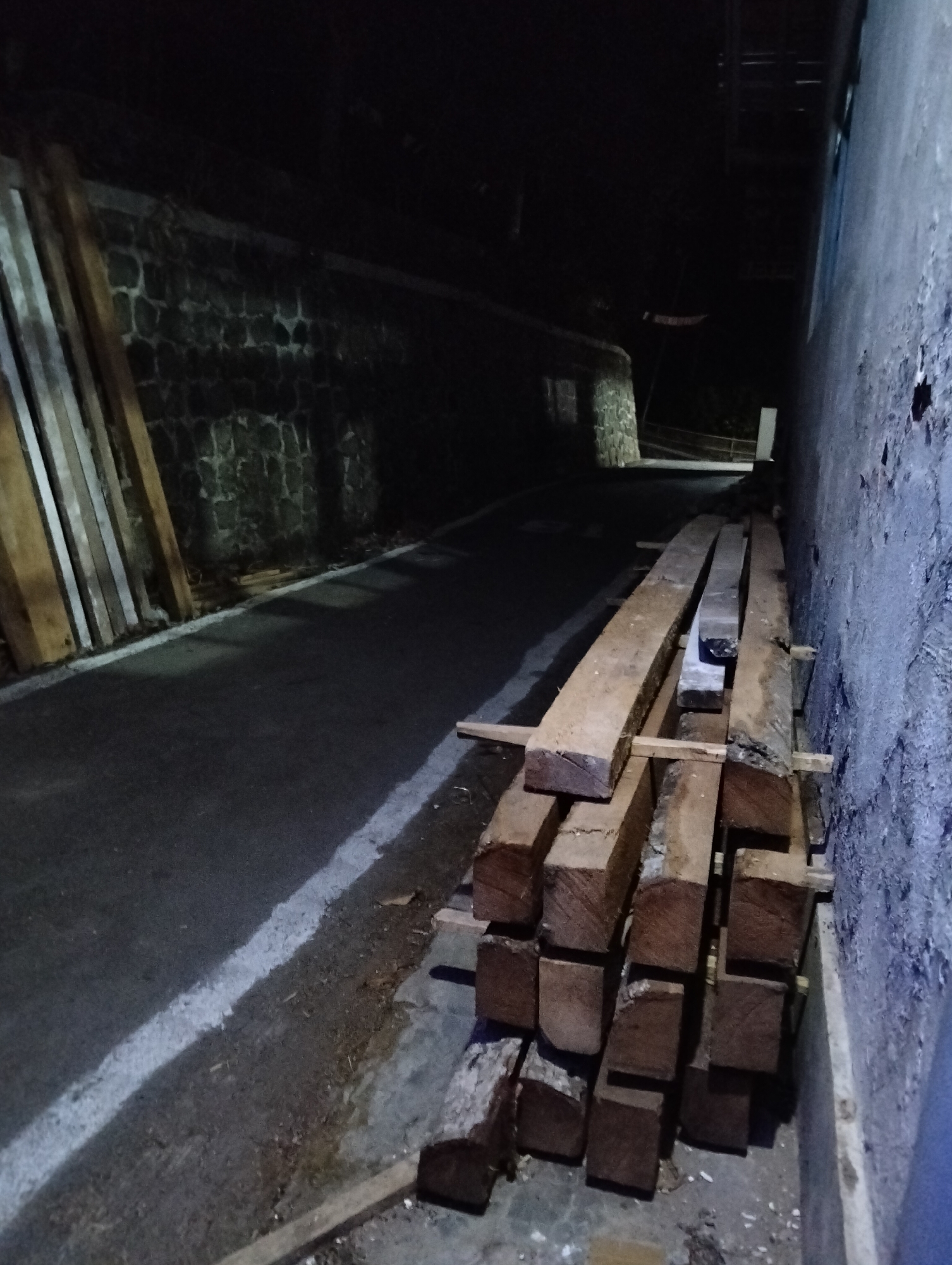 In the city of Paguyangan, Indonesia, a dark, asphalt road runs alongside a stone wall and a building. Several large, rough-hewn wooden beams are stacked on the side of the road, resting on smaller wooden supports. To the left of the road, more wooden planks are leaning against the stone wall. The scene is dimly lit, suggesting it is nighttime or a poorly illuminated area.
