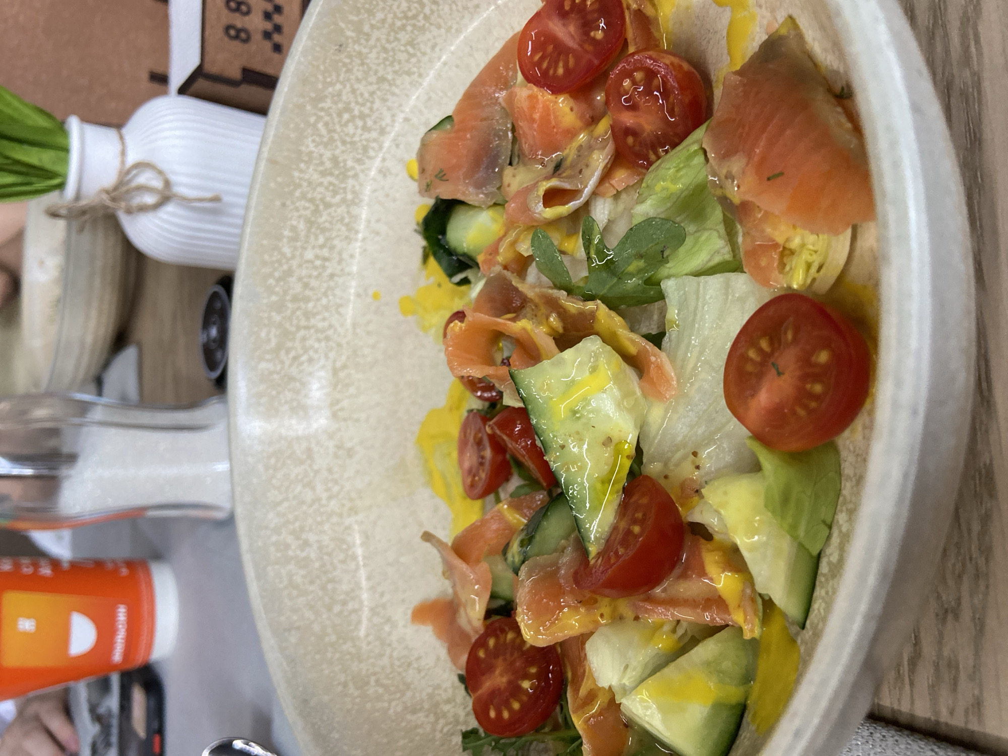 A close-up, overhead shot shows a vibrant salad in a speckled, cream-colored bowl. The salad is composed of fresh greens, sliced cherry tomatoes, thinly sliced cucumbers, and delicate pieces of smoked salmon. A light yellow dressing is drizzled over the ingredients. The background is blurred but suggests a restaurant setting with a red and white paper cup, a salt shaker, and a white vase with greenery. The overall scene depicts a prepared meal ready to be consumed. This image does not contain any information or context that would allow for the identification of its location in Tuymazy, Russia.