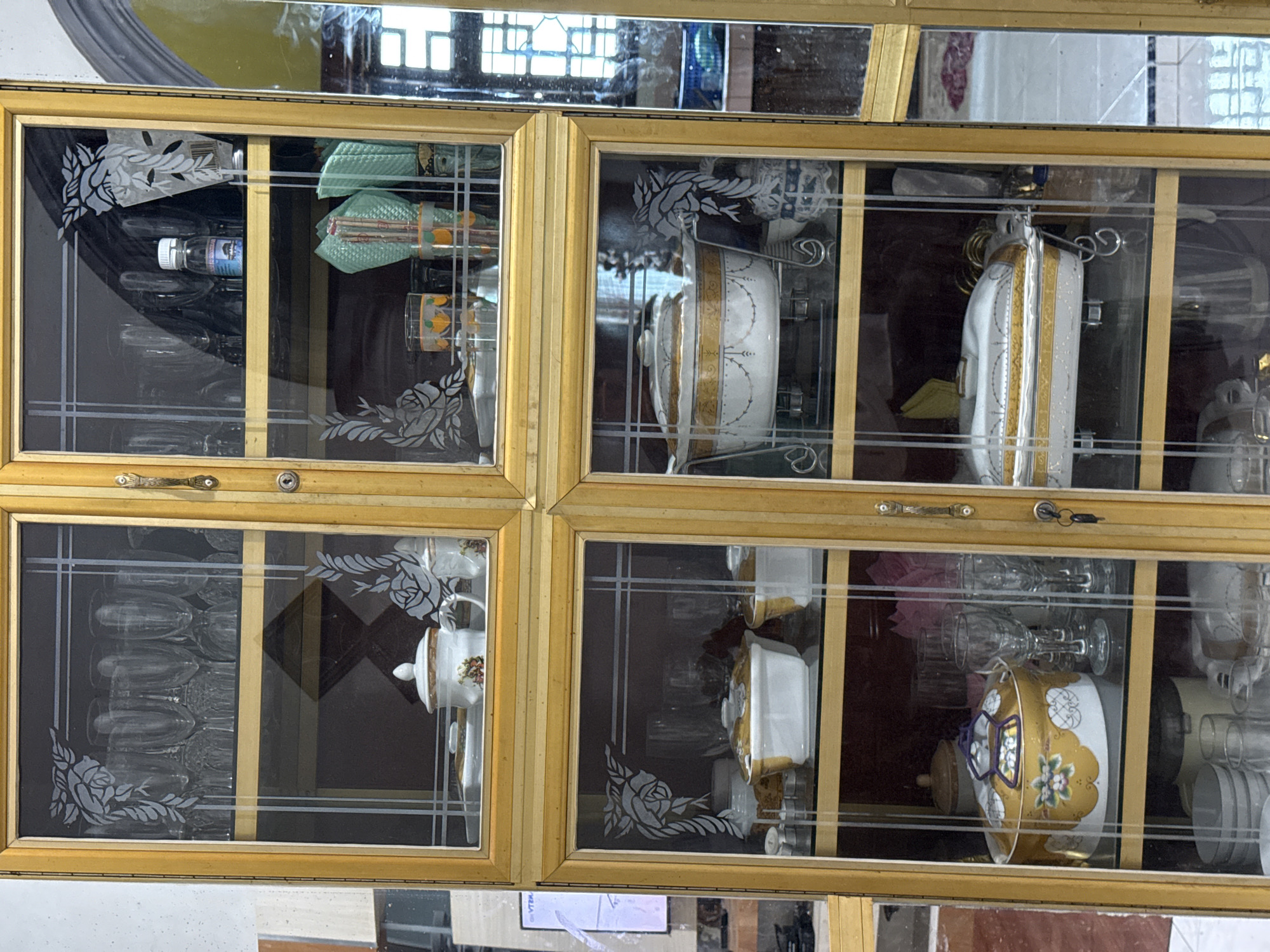 This image shows a display cabinet filled with glassware and dishes, likely in a home in Bireuenmeunasahblang, Indonesia. The cabinet is made of wood with a golden finish and features glass doors with decorative etched patterns.
The top left section of the cabinet contains several rows of clear stemmed glasses. The top right section holds more stemmed glasses, a small bottle of liquid with a blue label, and folded green napkins.
The middle left section displays two white ceramic serving dishes with gold trim. One dish is topped with a lid. Next to these dishes are some smaller white ceramic pieces and a dark brown lidded container.
The middle right section features a large white ceramic serving dish with an ornate gold pattern, resting on a metal stand. To the right of this dish, there is a small blue and white patterned ceramic item.
The bottom left section contains a large, round, yellow serving dish with a floral pattern and a blue lid handle. Several clear stemmed glasses are positioned to the right of this dish. Below this, stacks of white plates and more glassware are visible.
The bottom right section contains another white ceramic serving dish with a gold trim, also on a metal stand. To the right, a reflection in a mirrored surface shows a patterned wall and some objects.
The overall impression is a well-organized collection of dining ware displayed in a traditional cabinet.