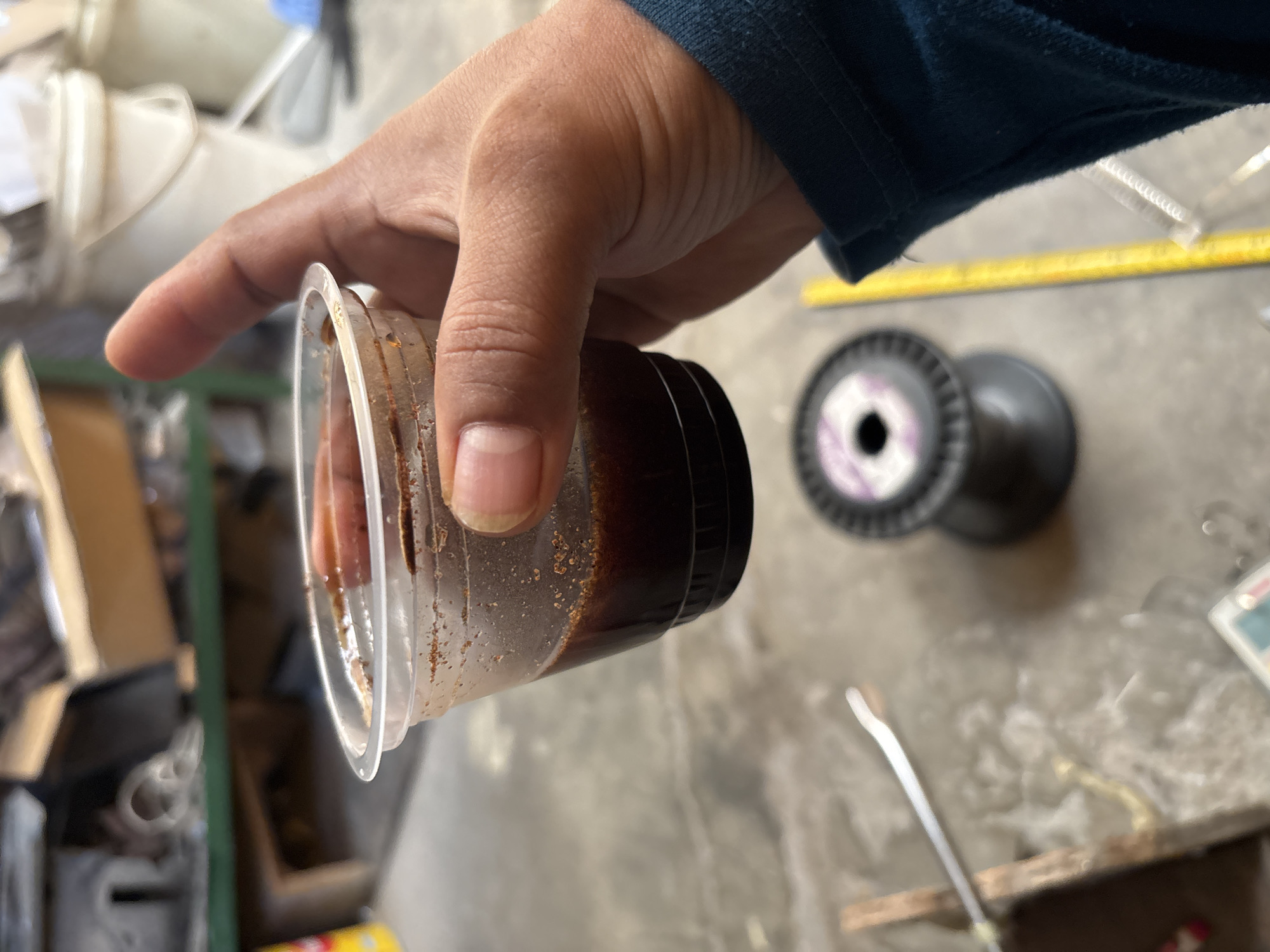 A person's hand holds a clear plastic cup filled with a dark liquid, possibly coffee or a similar beverage. The cup is slightly tilted, revealing sediment settled at the bottom. The background is blurred, but appears to be an outdoor or workshop environment with various objects and structures, including what looks like a wooden table with tools and a spool of wire. The image is taken from a close-up perspective, focusing on the cup and hand. The location is identified as Telagamurni, Indonesia.