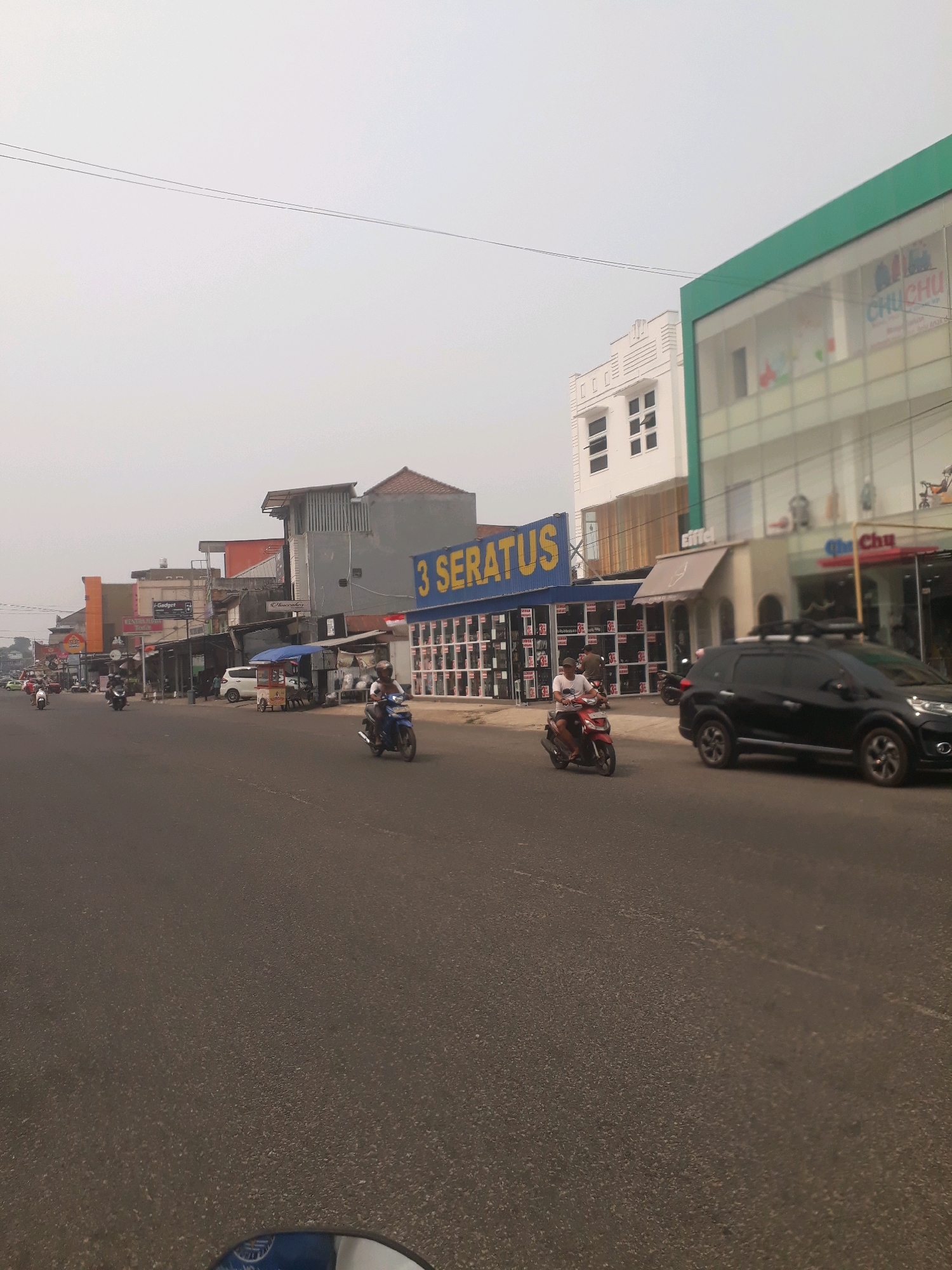 The image captures a street scene in Pangkalpinang, Indonesia, likely during daylight hours under an overcast sky. The road in the foreground is asphalt, with a motorcycle visible in the lower left corner, suggesting the photo was taken from another moving motorcycle. On the right side of the road, a black SUV is driving, and further down, a red motorcycle with a rider is also in motion. The buildings lining the street are varied in architecture. On the left, there are several commercial establishments with awnings and signs, including one with the text "I-Gadget." Further down the street on the left, a building with a red roof is visible. On the right side of the street, a prominent building with a green facade and the text "CHU CHU" is present. Adjacent to this is a building with the text "3 SERATUS" in large yellow letters, displaying numerous windows with goods inside. Another building with an awning labeled "Eiffel" is situated next to it. The overall impression is a busy commercial street with moderate traffic.