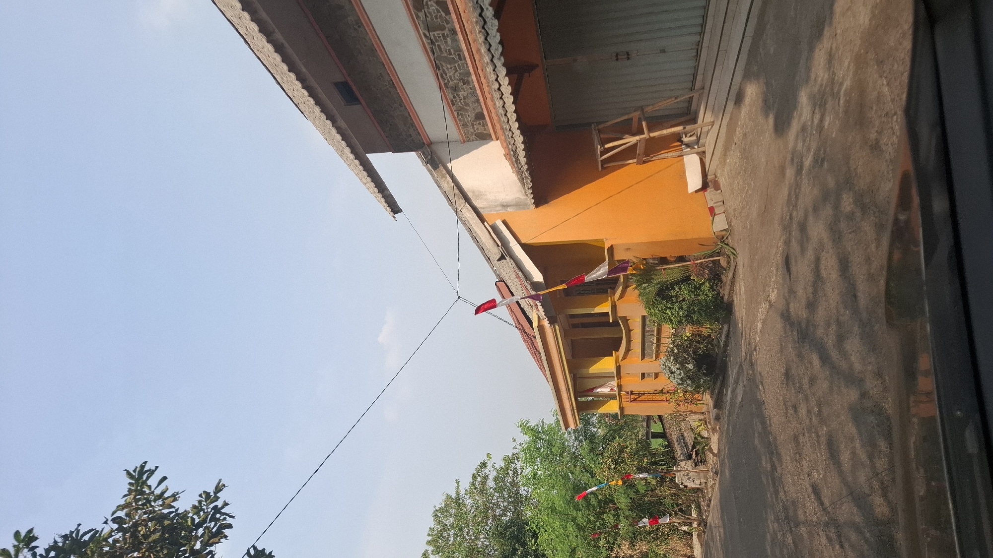 The image depicts a street scene in Cibinong Hilir, Indonesia, on a clear day. In the foreground, the dark, reflective surface of a car's dashboard or hood is visible, suggesting the photo was taken from inside a vehicle. The road ahead is paved and shows dappled sunlight and shadows cast by nearby trees and structures.
To the left of the road, a bright orange building with a covered porch area stands out. Several small Indonesian flags, featuring red and white stripes, are visible along the roadside and attached to the building's facade, indicating a festive or patriotic occasion. Dense green foliage and trees are present behind the flags and to the left of the orange building.
To the right of the orange building, another structure is partially visible. This building has a roof with grey, overlapping tiles and sections of stone-like cladding and lighter colored walls. A metal roll-up door is prominent on this section of the building. A wooden ladder-like structure stands against the orange wall near the roll-up door.
The sky is a clear, pale blue, with no visible clouds except for a faint wispy one in the distance. Overhead, electrical wires crisscross the scene. The overall impression is of a residential or commercial street in an Indonesian town.