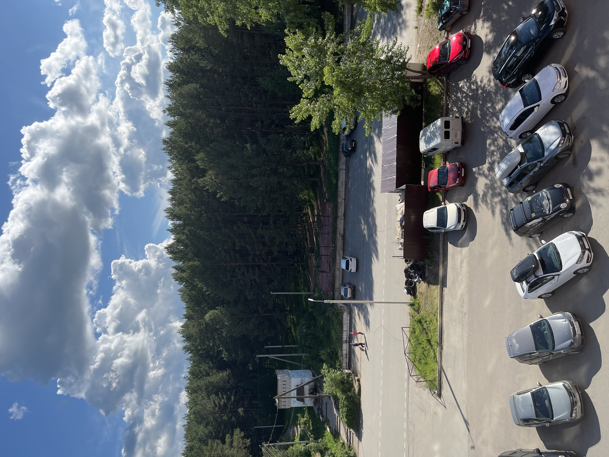 The image depicts a view from above, likely from a high-rise building, overlooking a street and parking area in the city of Chaikovsky, Russia. A dense forest lines the left side of the scene, contrasting with the urban environment on the right.
Several cars are parked along the right side of the street, varying in color and model. A red car, a white van, and several white and dark-colored sedans are visible.
A two-lane road with white lane markings runs through the center of the image. In the distance, two people appear to be walking or interacting near a lamp post. A small building or structure is visible among the trees closer to the bottom of the image, surrounded by overgrown vegetation and electrical poles.
The sky features a mix of blue and cumulus clouds, suggesting a sunny day.