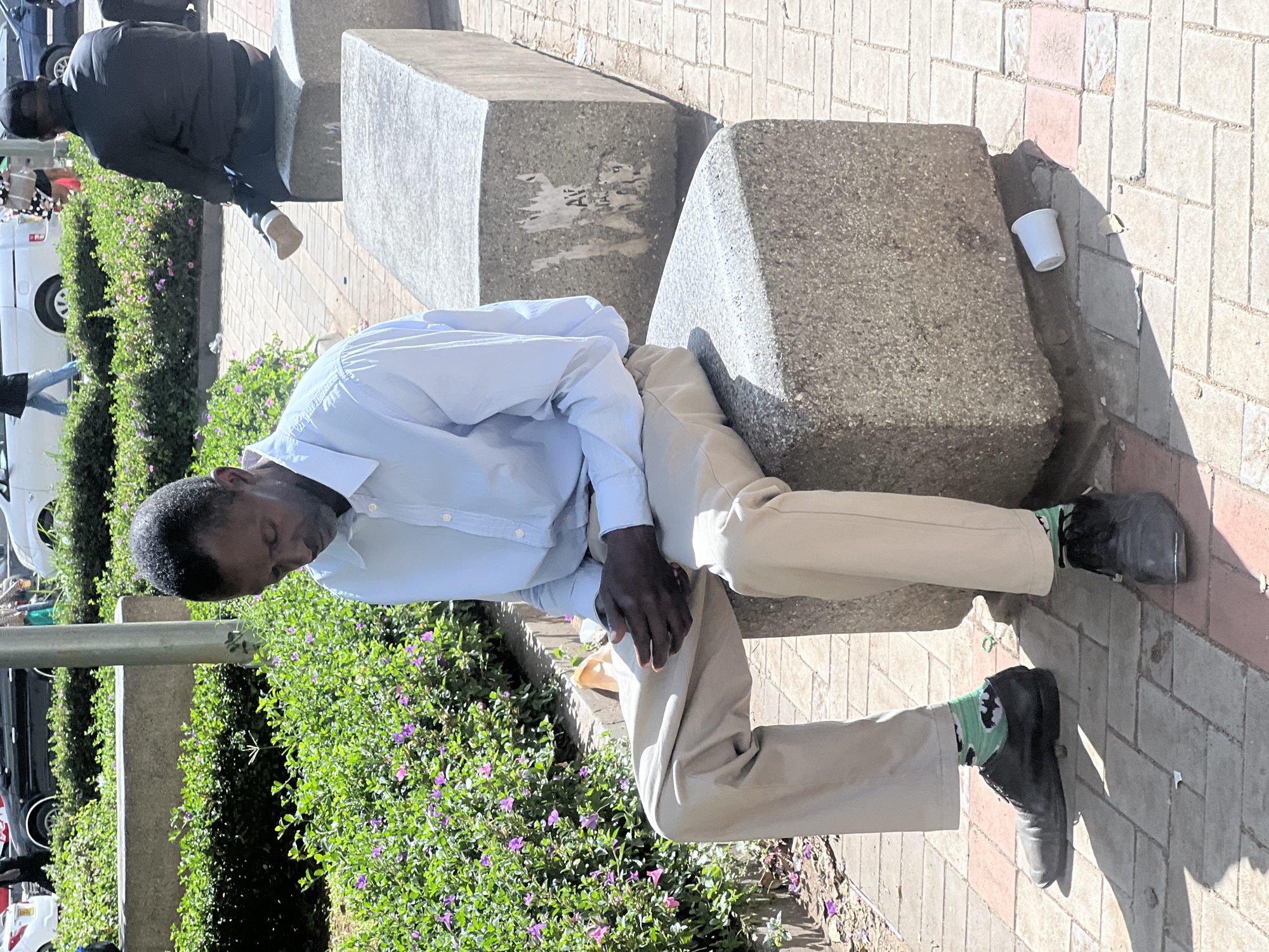 A man in a light blue shirt and khaki pants is sitting on a series of grey, concrete blocks. He appears to be sleeping. Another man is sitting on a block behind him, also appearing to rest. A white cup sits on top of one of the concrete blocks. There are green bushes with small purple flowers next to the concrete blocks. The scene is located on a brick sidewalk, with parked cars and buildings visible in the background. This photograph depicts a public space in Nairobi, Kenya.