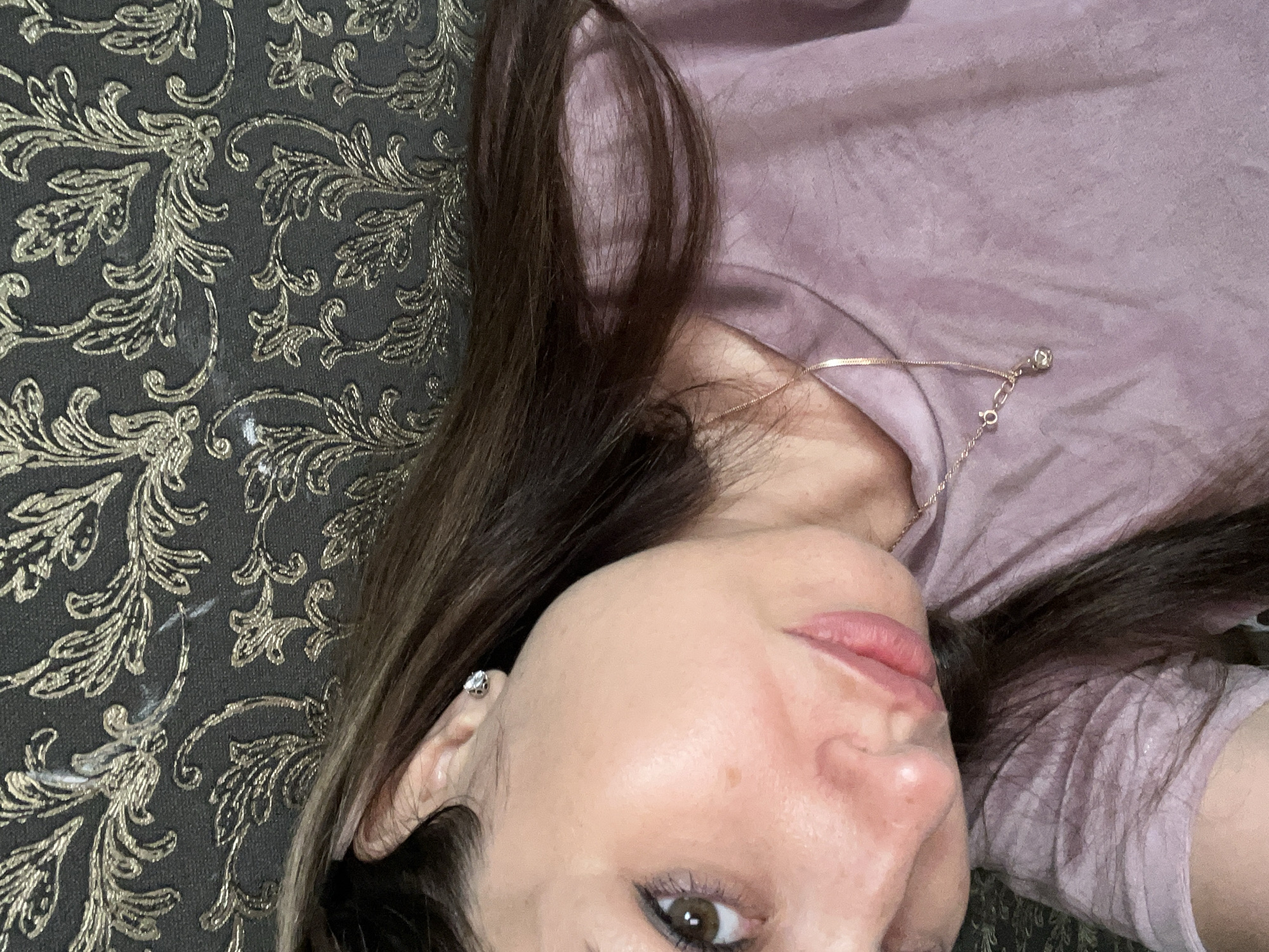 A woman is looking at the camera, her head tilted to the side. She has long, dark brown hair and brown eyes. She is wearing a light purple t-shirt and a delicate gold necklace with a small pendant. A diamond stud earring is visible in her left ear. The background is a dark gray fabric with a gold floral pattern. The image appears to be a selfie, taken from a close-up perspective.