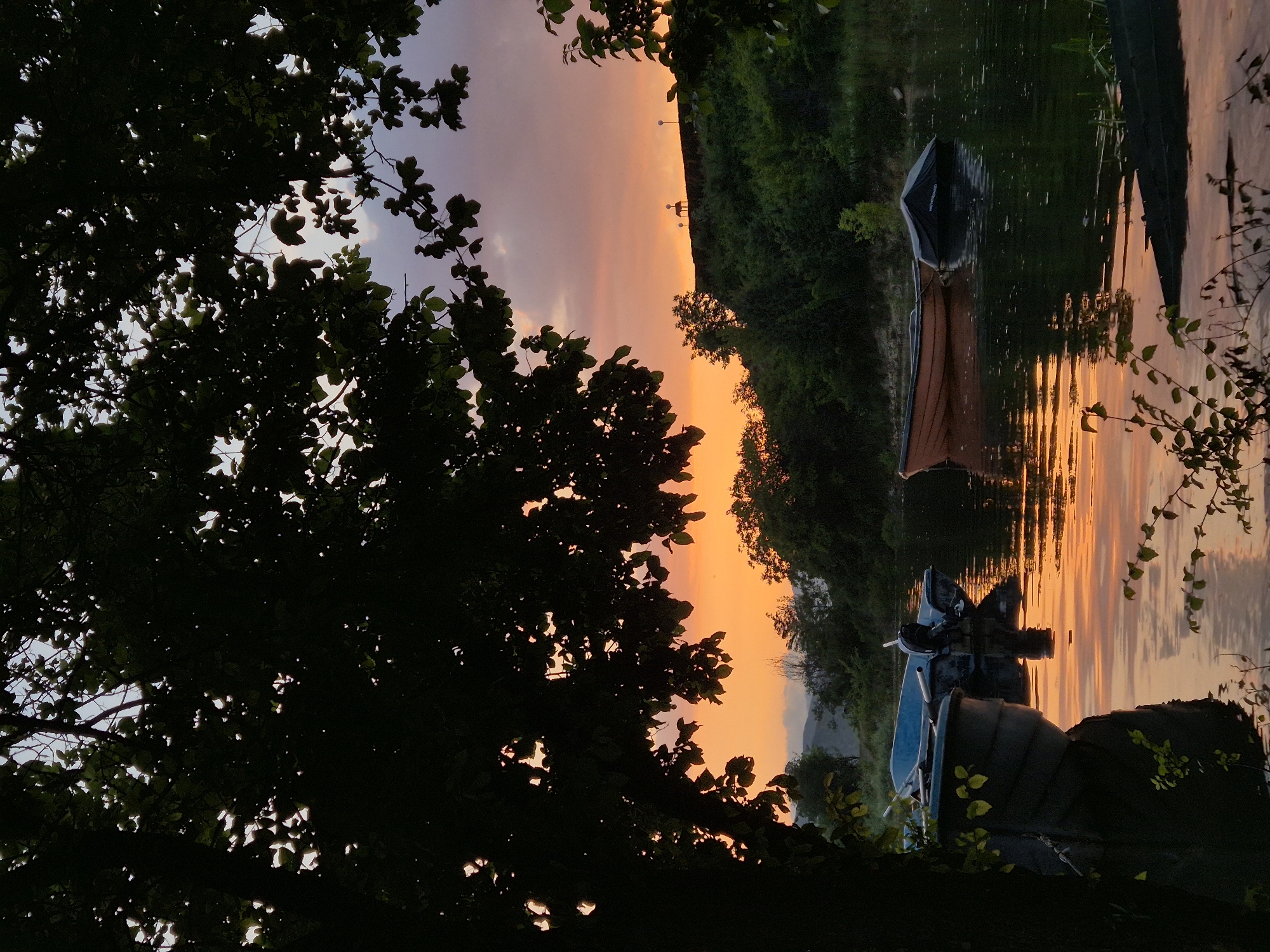 This image captures a serene sunset view over a tranquil body of water, likely a lake or a wide river, in Glavatartsi, Bulgaria. The photograph is taken from a vantage point offering a partial glimpse through dense, dark foliage, which heavily frames the left side and foreground of the scene.
The sky, dominating the upper half, displays a stunning gradient of colors characteristic of twilight. It transitions from soft lavender and muted grey-blue at the top left, where faint clouds are visible, to an intense, fiery orange and golden hue stretching across the horizon.
The calm surface of the water, occupying the lower and right portions of the frame, perfectly reflects the vibrant sunset. This creates a mesmerizing pattern of shimmering orange and yellow streaks that extend across the water, mirroring the sky above.
Two boats are visible on the water, seemingly moored close to the leafy bank. The boat in the upper right part of the water has a brownish-orange hull with a dark canopy or cover; faint, illegible text might be present on this cover. Another boat, darker in color (possibly blue or black) and equipped with some apparatus, is positioned further to the right.
The immediate foreground is characterized by the dark, silhouetted shapes of tree branches and numerous leaves, which provide a natural frame for the dramatic sunset. These trees appear to be very close to the viewer, suggesting a perspective from a wooded shore.
In the far background, across the water, a dark, undulating landmass, possibly a forested hill or embankment, is silhouetted against the bright sky. Small, indistinct structures, possibly lampposts, can be faintly discerned along its crest.
The overall impression is one of peaceful natural beauty at dusk. No people are visible, emphasizing the quiet solitude of the moment. The scene evokes a tranquil and contemplative mood.