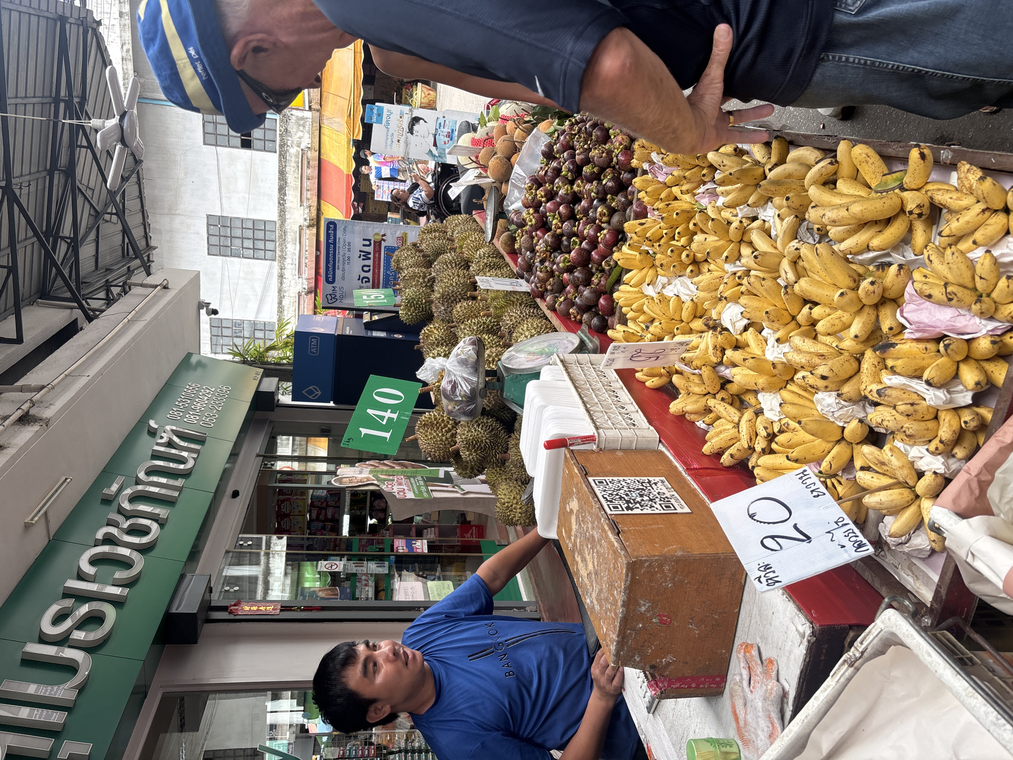 This is a medium shot of a fruit stand in Chiang Mai City Municipality, Thailand. A male vendor, wearing a blue t-shirt with "Bangkok" written on it, stands behind the stall. He is looking to his left, speaking to a customer who is partially visible on the right side of the frame, wearing a blue and yellow hat and sunglasses. The stall is laden with a variety of fruits, including durian, mangosteen, and numerous bunches of bananas. A sign in front of the bananas indicates a price of "20". In the background, a store with a green awning and Thai lettering is visible, along with other market stalls and buildings. The overall scene depicts a bustling outdoor market environment.