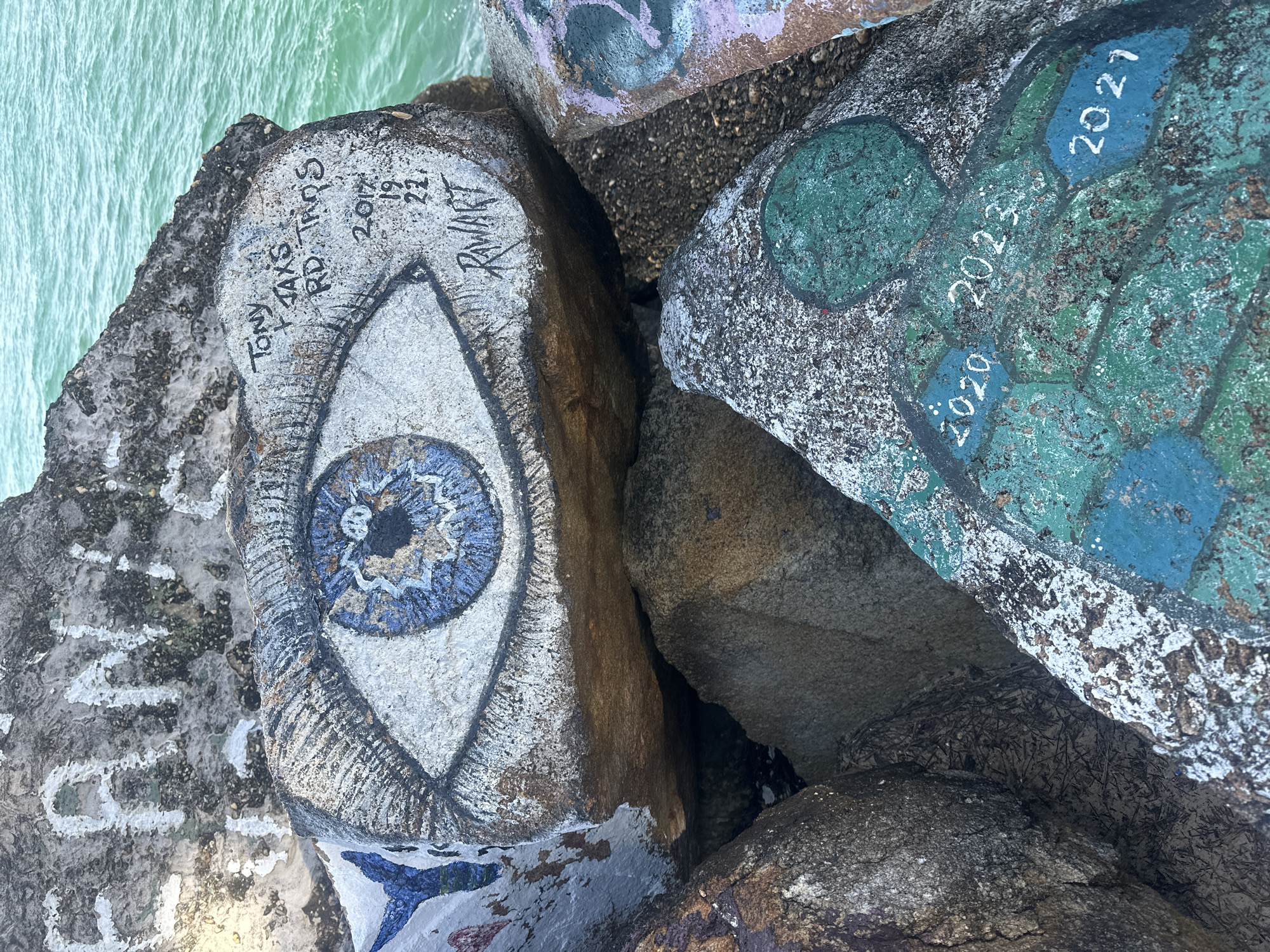 The image shows a close-up of graffiti art on rocks near a body of water. One large rock features a painted eye with detailed blue and black iris. Above the eye, graffiti in black marker includes the words "TONY", "TAXES", and "RD TRQS", along with dates "2017", "19", "22", and "RAWART". Another rock in the foreground has a painted turtle-like shape in green and blue, with the years "2020", "2023", and "2021" written in white. The background shows the turquoise water of the ocean. The context of Nambucca Heads, Australia, is not visible in the image.