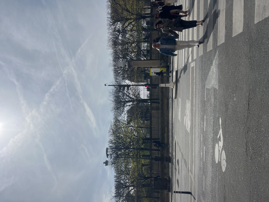 The image captures a street scene in Paris, France, bordering what appears to be a formal garden or park, consistent with the perimeter of the Tuileries Garden. The sky is bright and sunny, featuring a light blue hue with several white contrails.
In the foreground, an asphalt road displays distinct white markings, including a pedestrian zebra crossing, a bicycle lane symbol, and directional arrows. A black bollard is visible on the far left of the road.
The midground features two main subjects walking hand-in-hand across the pedestrian crossing from right to left. The male subject is bald, wearing sunglasses, a light blue denim jacket over a light-colored top, and off-white trousers. The female subject has dark hair, wears sunglasses, a dark jacket, dark trousers, and carries a black shoulder bag. Additional pedestrians are partially visible further to the right on the crossing. A black pole supports a red traffic light, situated behind a white base, on the right side of the frame. A bicycle is parked near this structure. On the left side of the road, a dark, ornate lamppost with two light fixtures stands near a low wall. Another individual is leaning against this wall further back.
The background is dominated by a tall black wrought-iron fence, separating the street from the park or garden. Behind the fence, a dense row of deciduous trees displays early spring foliage with visible green buds. A classical white stone urn on a pedestal is positioned prominently among the trees, visible above the fence line to the right of the ornate lamppost.