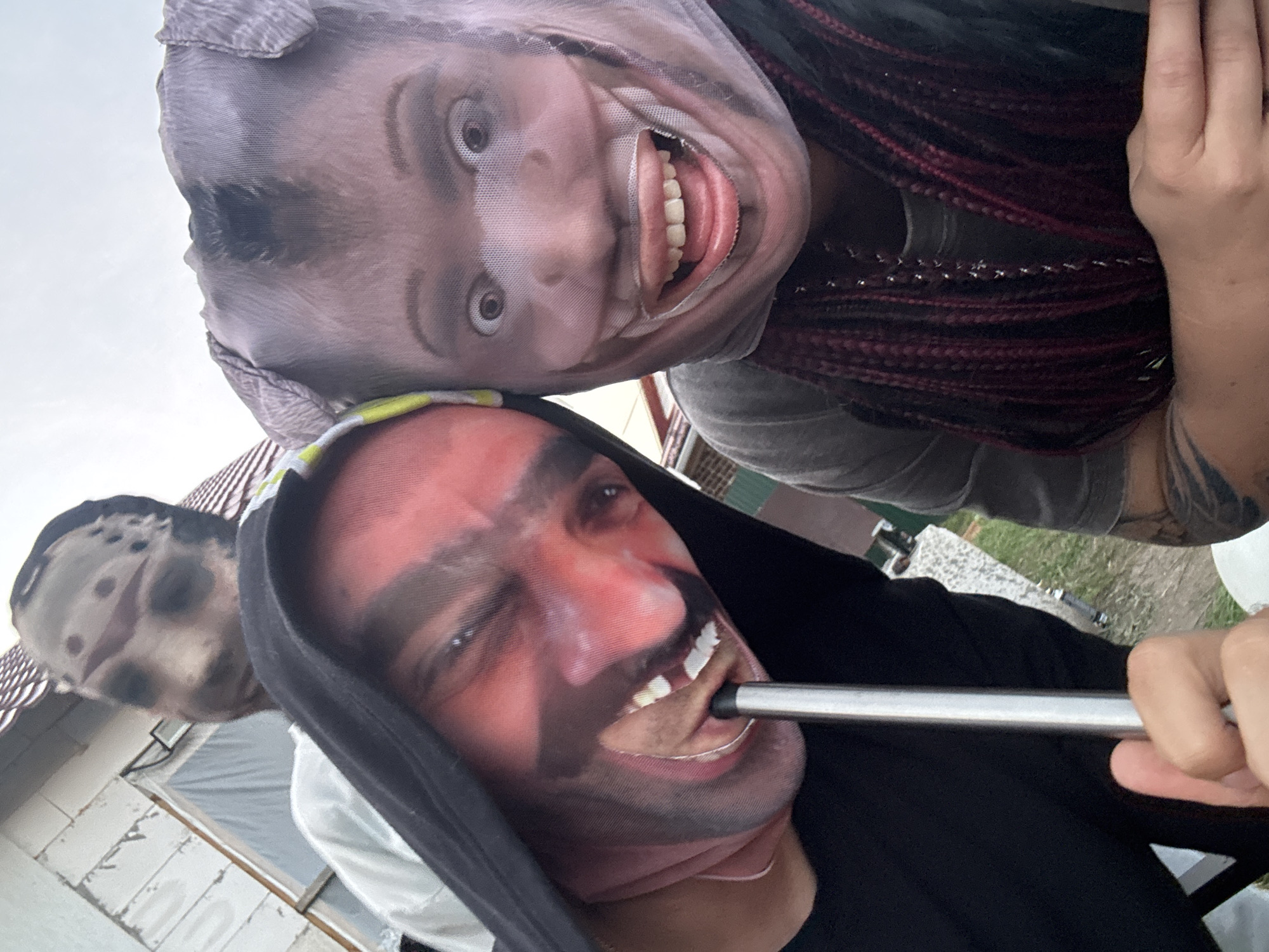 Two individuals are posing for a selfie outdoors, their faces covered by novelty masks. The person on the left wears a mask with a red, devilish face and a black hood. They are holding a silver cylindrical object, possibly a straw, in their mouth. To the right, another person wears a mask resembling a pig or similar animal face with wide, open eyes and a large, grinning mouth. This person has dark, braided hair with red streaks. In the background, partially obscured, is a third individual wearing a mask that appears to be a horror-themed mask, possibly of a character like Jason Voorhees from the Friday the 13th movies. The setting appears to be a yard with a partially constructed building with exposed framing and siding. There is grass and gravel on the ground. The sky is overcast. There is no visible indication of the specific city of Узюково, Russia, in the image.