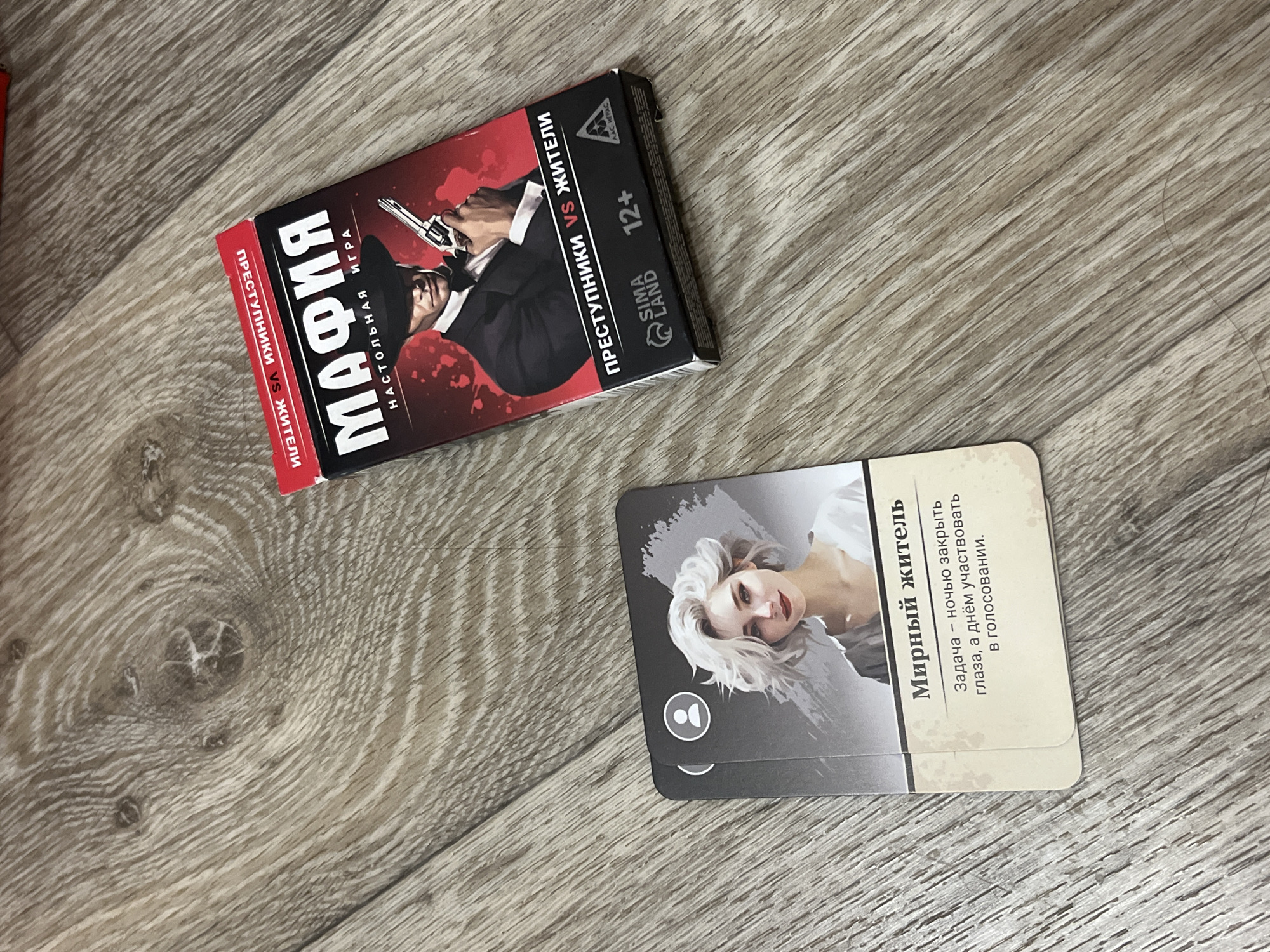 The image shows two game cards and a box for the board game "Mafia". The box is red and black and features an illustration of a man in a suit holding a gun. The cards are laid out on a wooden surface. The top card has an illustration of a woman with white hair and the text "Мирный житель" (Peaceful Resident). The bottom card is partially visible and appears to be another game card. The background is a wooden floor. There are no discernible landmarks or specific locations within the city of Дачное, Russia, visible in this image.