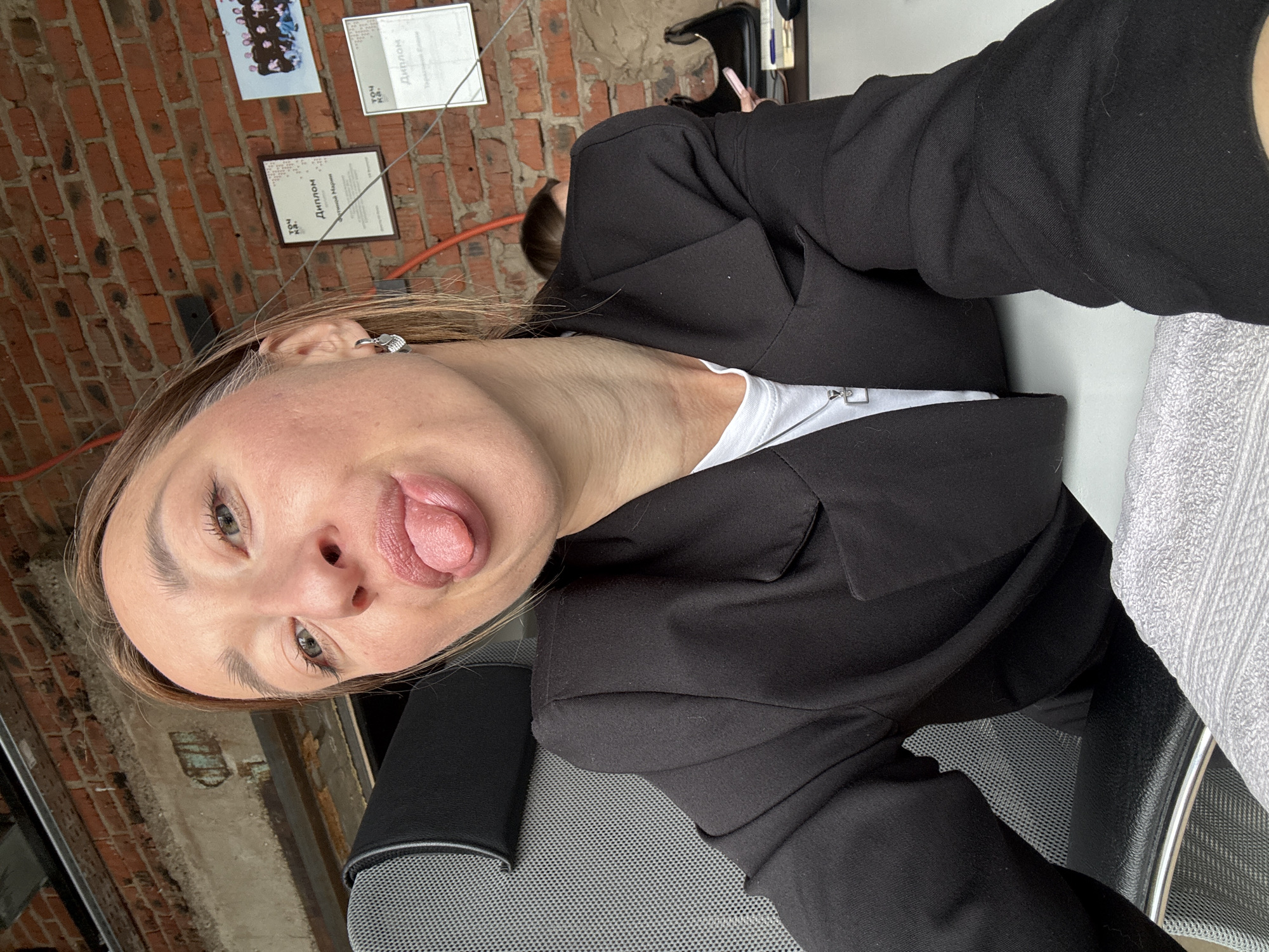 A person is sitting in a black office chair, wearing a black blazer over a white shirt. The individual has light skin, light brown hair, and is sticking their tongue out at the camera. The background is a brick wall with framed documents hung on it. The location is Chaikovsky, Russia, based on the language on the framed documents. The person is at a desk with a white surface. There is another person in the background.