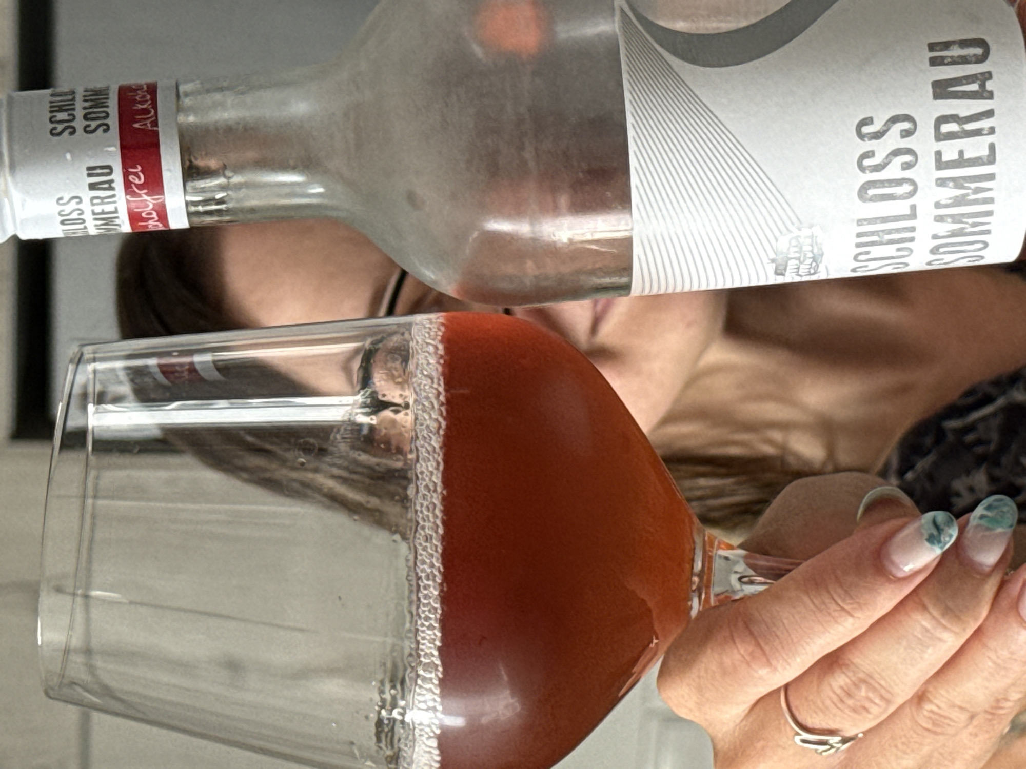 A person, partially obscured by a glass of reddish-brown liquid and a bottle, is in the foreground. The person is holding the glass with their left hand, which is adorned with a ring. The glass contains a frothy liquid. Beside the glass is a bottle with a white label featuring the text "SCHLOSS SOMMERAU ALKOHOLFREI" in black and red lettering. The background is blurred, suggesting an indoor setting. There are no discernible details about the location in Mesogi, Cyprus.