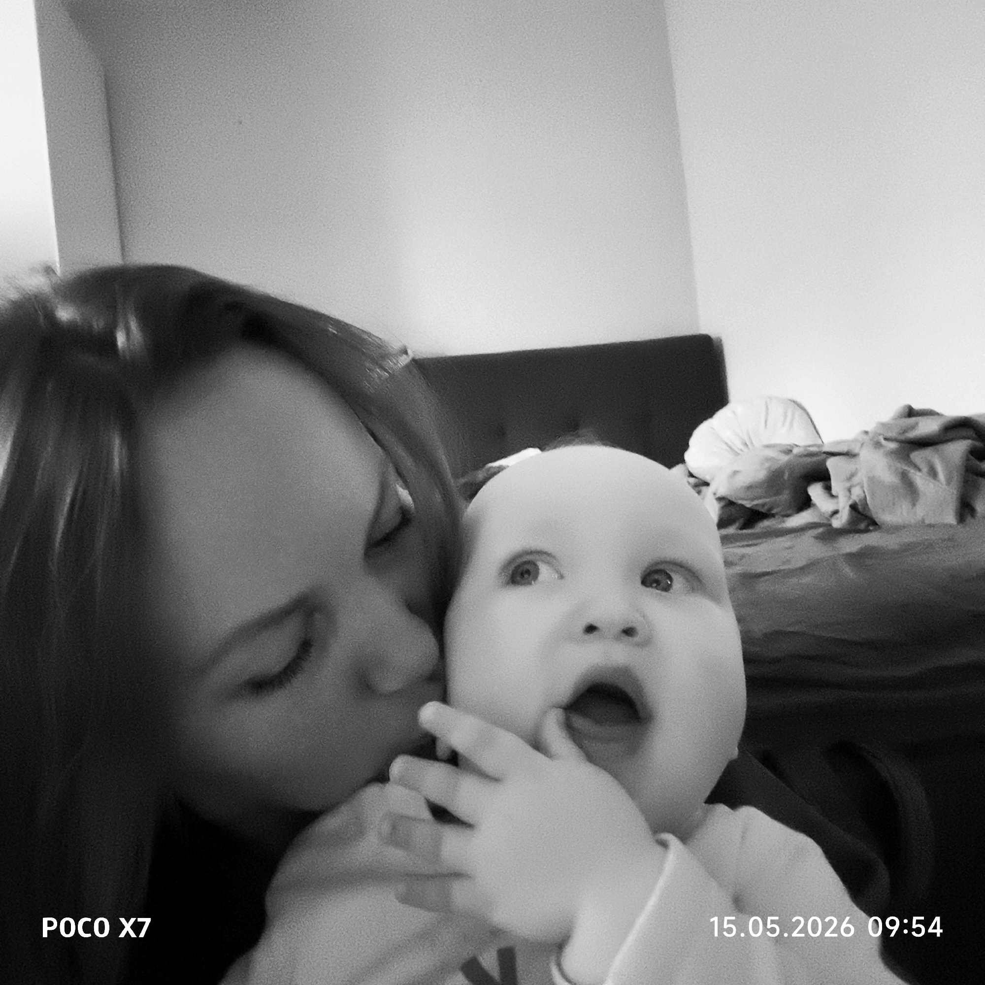 The black and white photograph shows a woman with dark hair kissing a baby on the cheek. The baby is looking upwards with an open mouth and a hand near its face. The background includes a couch with a pillow and draped cloth. The lower left corner of the image displays the text "POCO X7," and the lower right corner shows "15.05.2026 09:54." The photograph's setting appears to be indoors, possibly in a domestic environment. The photograph does not confirm if this image was taken in Chaikovsky, Russia, nor provides any specific information about it.