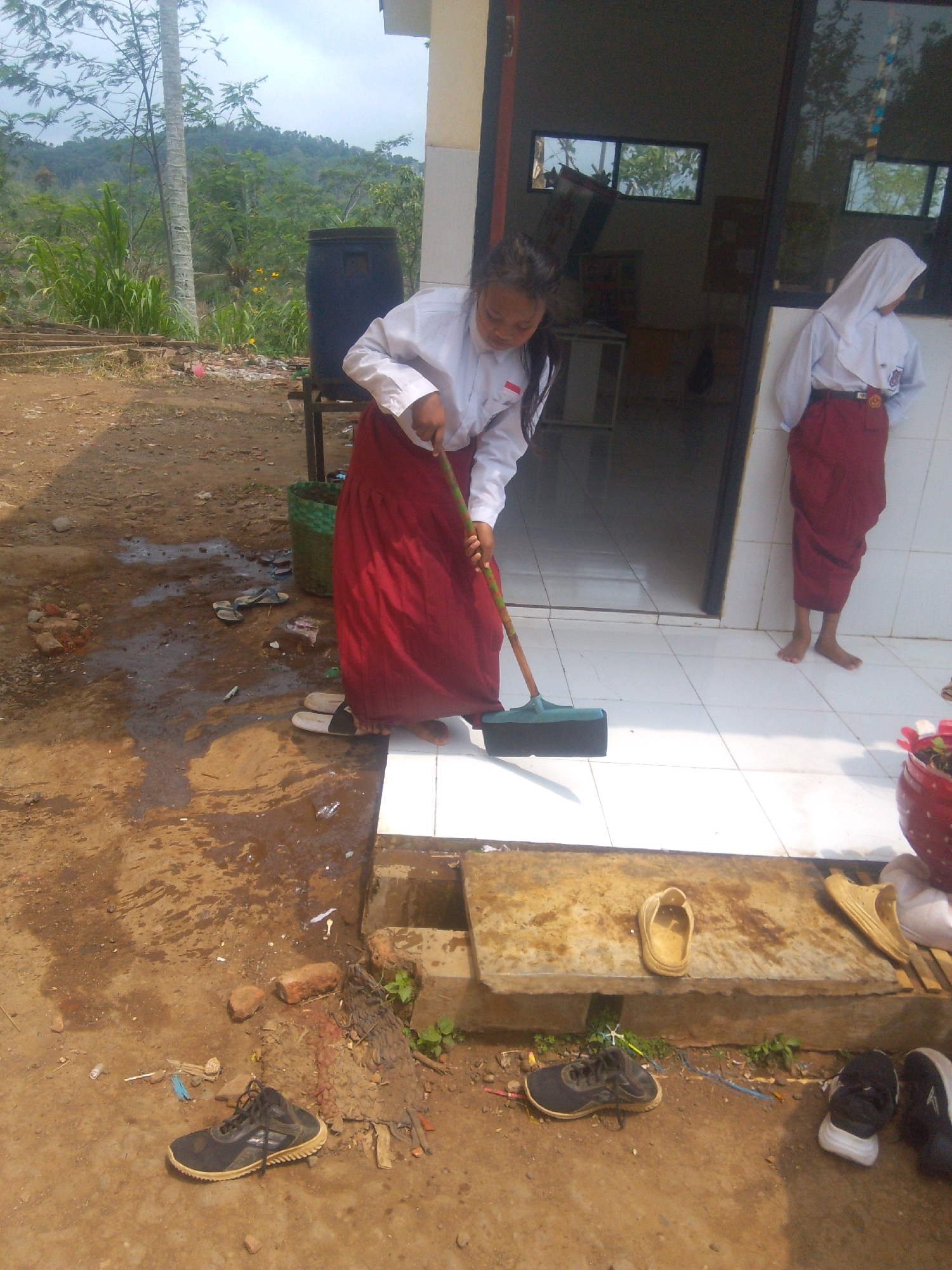 A young female student, wearing a white long-sleeved shirt and a red skirt, is sweeping the tiled floor outside a building with a green-handled broom. Another female student, also in a white shirt and red skirt and wearing a headscarf, stands to the right, facing away from the camera. The ground outside the building is a mix of dirt, water puddles, and scattered debris. Several pairs of shoes and sandals are visible on the ground and on a concrete ledge. In the background, there is lush green vegetation, trees, and a hill under a cloudy sky, suggesting a rural or semi-rural setting. The building appears to be a school or similar institution.