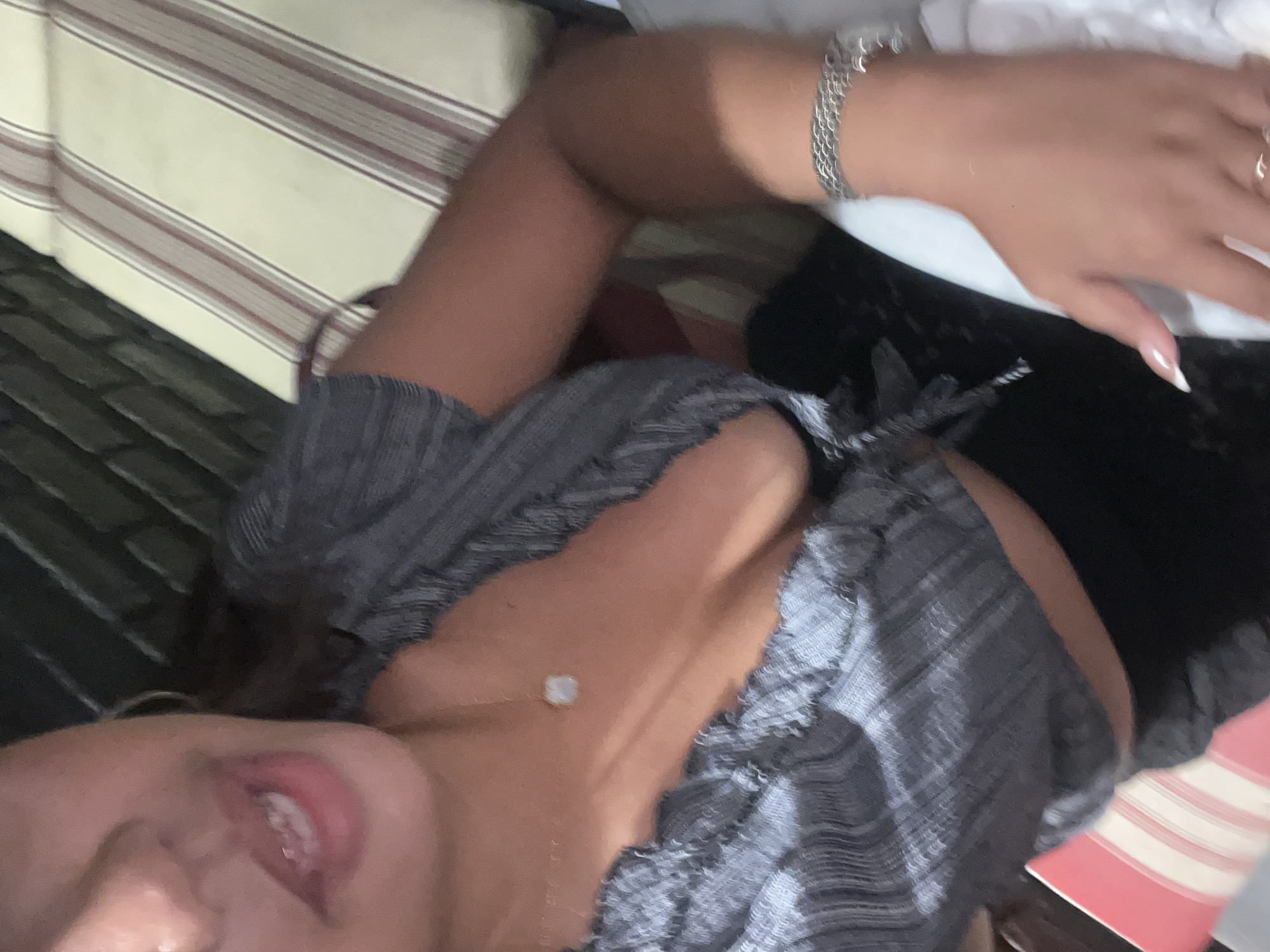 The image shows a close-up, tilted view of a person's upper body. The subject is wearing a grey, ruffled top and has a light-colored necklace with a small, square pendant. A silver bracelet is visible on their left wrist, and a ring is on their right index finger. Their fingernails are long and painted white. The person's mouth is slightly open, and their lips appear to be red. A portion of what looks like a striped cushion or fabric is visible in the background. The scene suggests a relaxed, possibly indoor setting. The lighting appears to be artificial, casting some shadows and highlighting the subject. There is no visible text in the image.