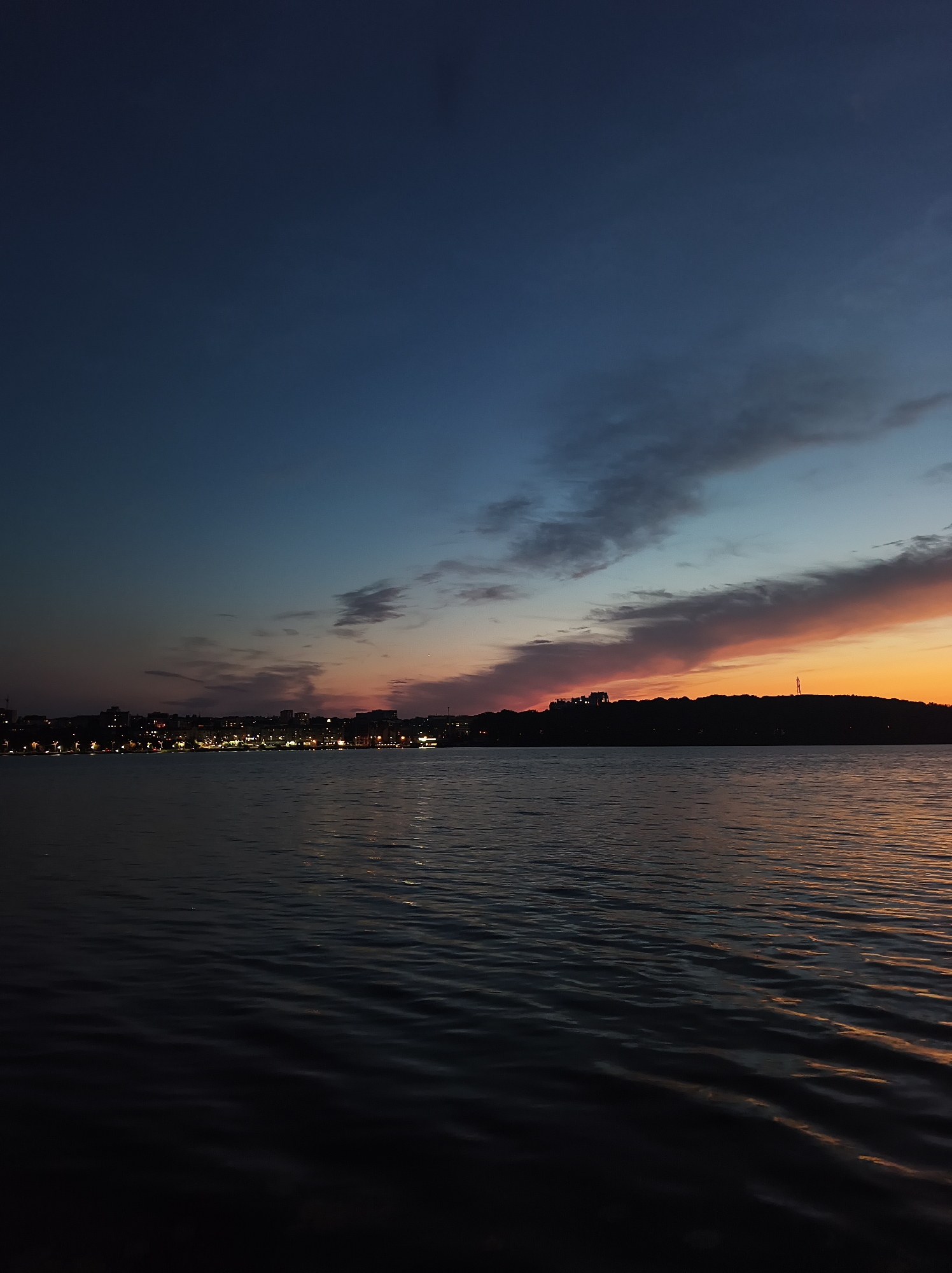 The image depicts a body of water, likely Ternopil Lake, at dusk. The sky is a gradient of deep blue at the top transitioning to hues of orange and pink near the horizon, indicative of a sunset. Wispy clouds are scattered across the sky, catching the fading light. Silhouetted against the brighter sky is a cityscape on the left side of the frame, with numerous lights indicating buildings and a populated area. On the right side, a darker, tree-lined silhouette of land is visible with a solitary tower-like structure on the horizon. The water in the foreground is dark and rippled, reflecting the colors of the sky. No people or distinct actions are visible, creating a serene and atmospheric scene of the urban landscape by the water at twilight.