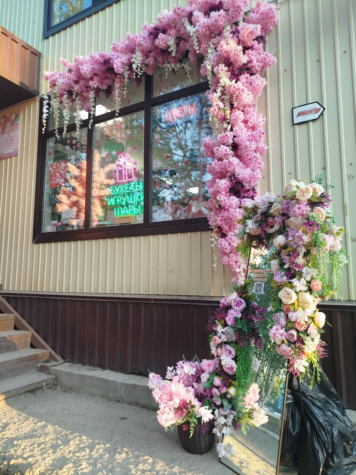 The image depicts the exterior of a flower shop in the city of Mirny, Russia, adorned with abundant pink floral decorations. The shop's facade features a large window displaying neon signs. One sign, in green neon, reads "БУКЕТЫ ИГРУШКИ ШАРЫ," which translates to "BOUQUETS TOYS BALLOONS." Another sign with a gift box icon is also visible. The window is framed by elaborate garlands of artificial pink and white flowers, with some cascading downwards.
To the right of the entrance, a white sign with a black arrow points to the right and says "Мицелия" (Mitselia), indicating the direction of the business. In front of the shop, large floral arrangements are displayed, including a tall arrangement of pink and cream-colored flowers and greenery to the right, and a basket of pink flowers to the left. A large black trash bag is positioned behind the tall floral arrangement. The shop appears to be situated on a street with a concrete path leading to stairs on the left side of the image. The ground is covered with sand and gravel, with dappled sunlight casting shadows. The building's exterior is clad in beige corrugated metal siding, with a brown corrugated metal base. The overall impression is a vibrant and festive storefront designed to attract customers to the flower shop.