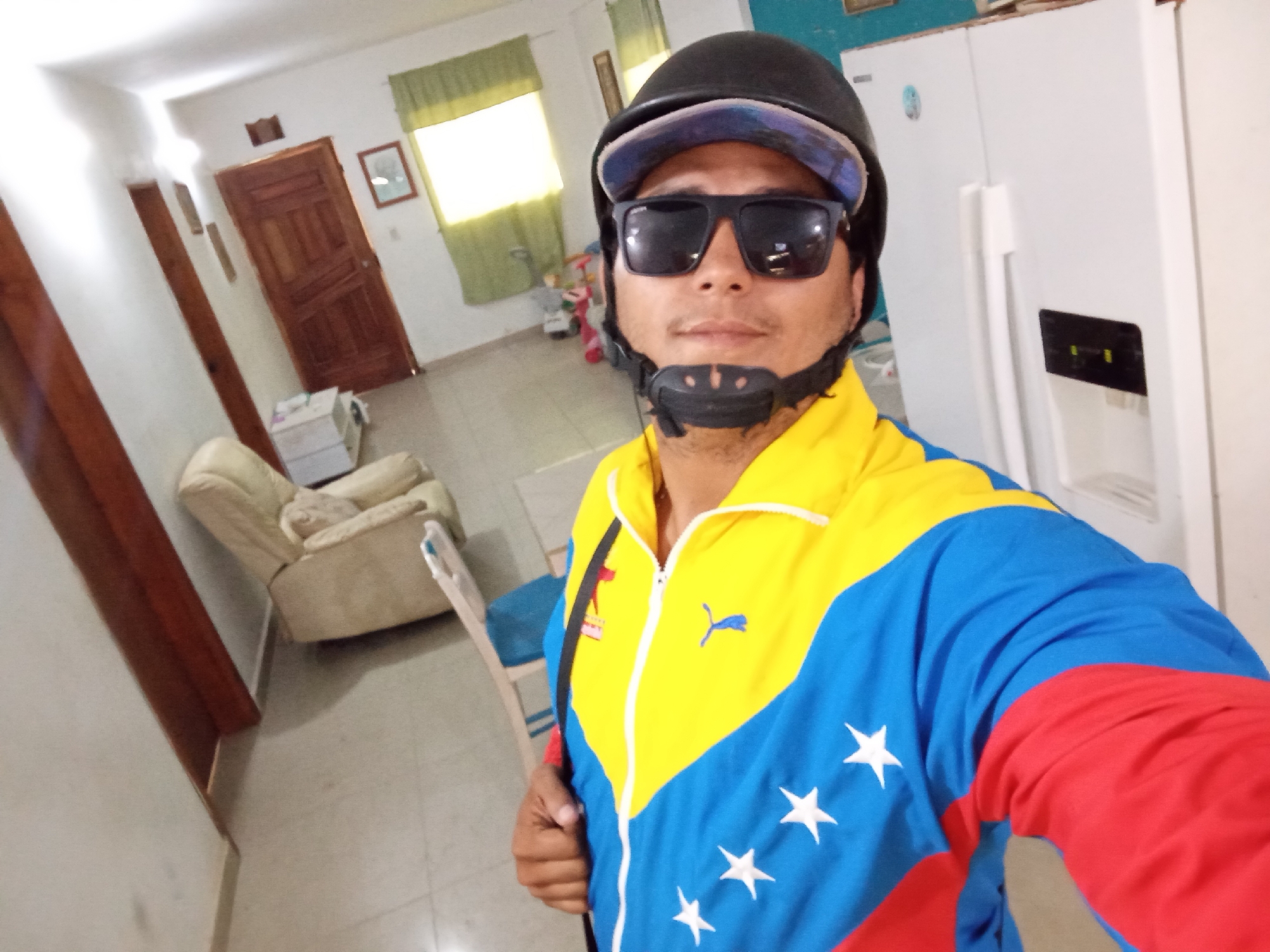 A man wearing a black helmet, sunglasses, and a jacket with the colors and stars of the Venezuelan flag is standing indoors. The jacket has yellow, blue, and red sections, with white stars on the blue part. Behind him is a white refrigerator, and in the background, there's a living room area with a beige armchair, a wooden door, and a window with green curtains. The location is in a house interior, potentially in or near the city of Juan Griego, Venezuela, due to the patriotic clothing.