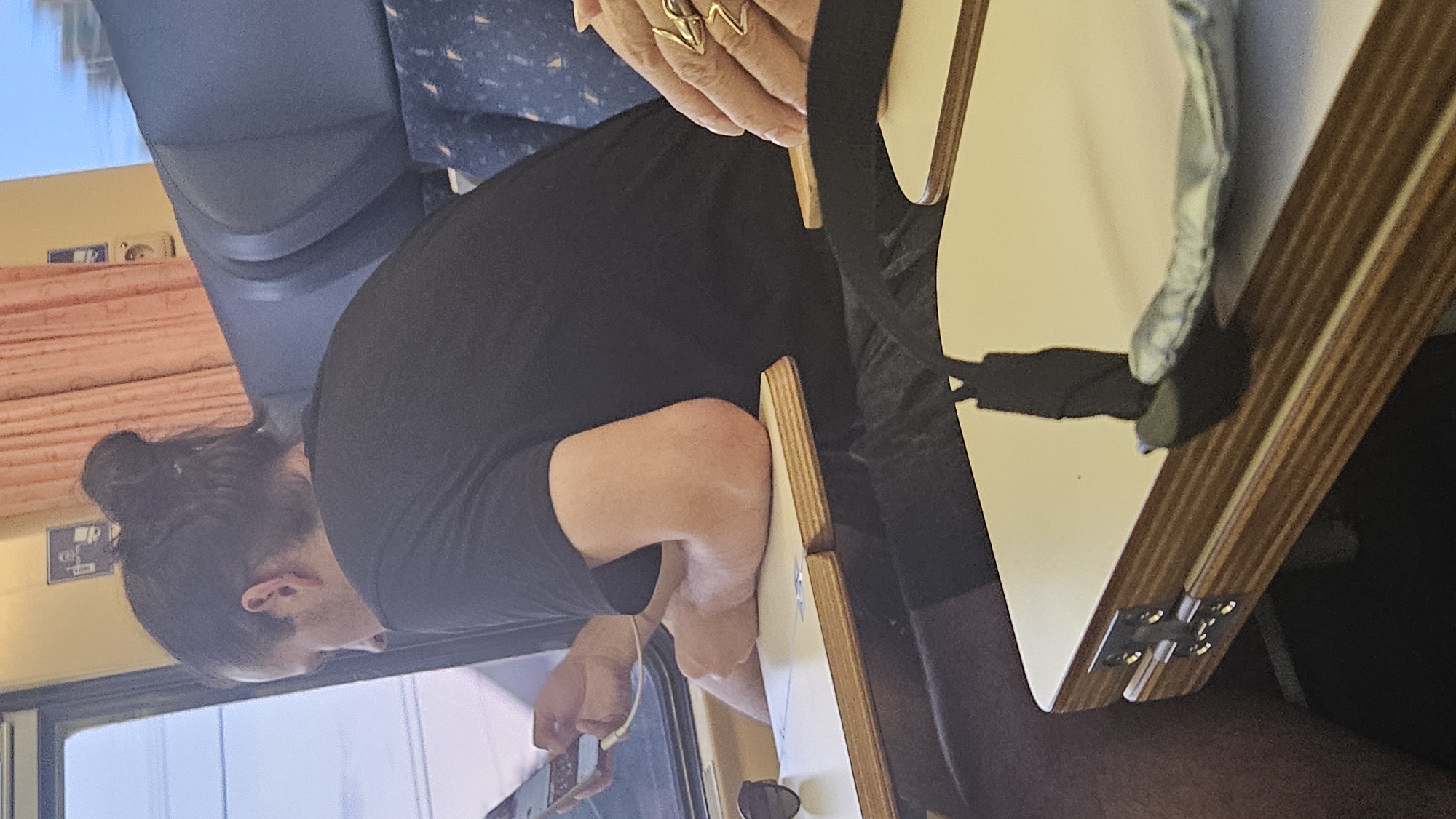 The image captures a person with dark hair styled in a bun, wearing a black t-shirt, seated inside what appears to be a train. The person is looking down at a mobile device, possibly a smartphone, connected to earphones. They are positioned next to a window, with a tray table in front of them. On the tray table, a pair of sunglasses is visible. Another person's hand, adorned with prominent rings, is also partially visible in the foreground, resting on another tray table. The interior of the train features blue seats with a patterned design and peach-colored curtains. The lighting suggests it is daytime, with light filtering in from the window. The background shows a blurry view of the outside, indicating the train is in motion. There is no discernible information in the image that would place this scene in the city of Olcnava, Slovakia.