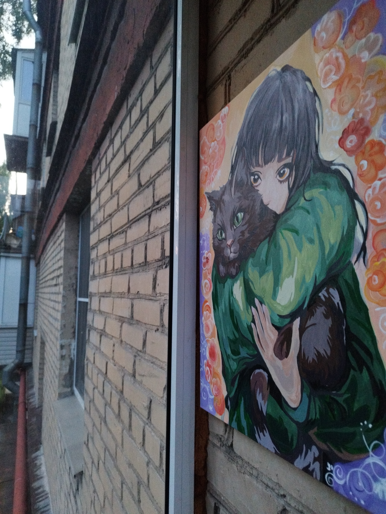 A painting of a young anime-style woman with dark hair and large amber eyes embraces a gray cat. The woman is wearing a green garment, and the cat's green eyes are wide open. The background of the painting features abstract floral shapes in shades of orange, pink, and purple. The painting is mounted on a brick wall, and the scene is viewed through a window. To the left of the painting, a brick building with a red trim and dark windows is visible, with a drainpipe running vertically down the side of the building. The overall scene suggests an outdoor setting, possibly a courtyard or alleyway in Saint Petersburg, Russia, given the architectural style of the building and the brickwork.