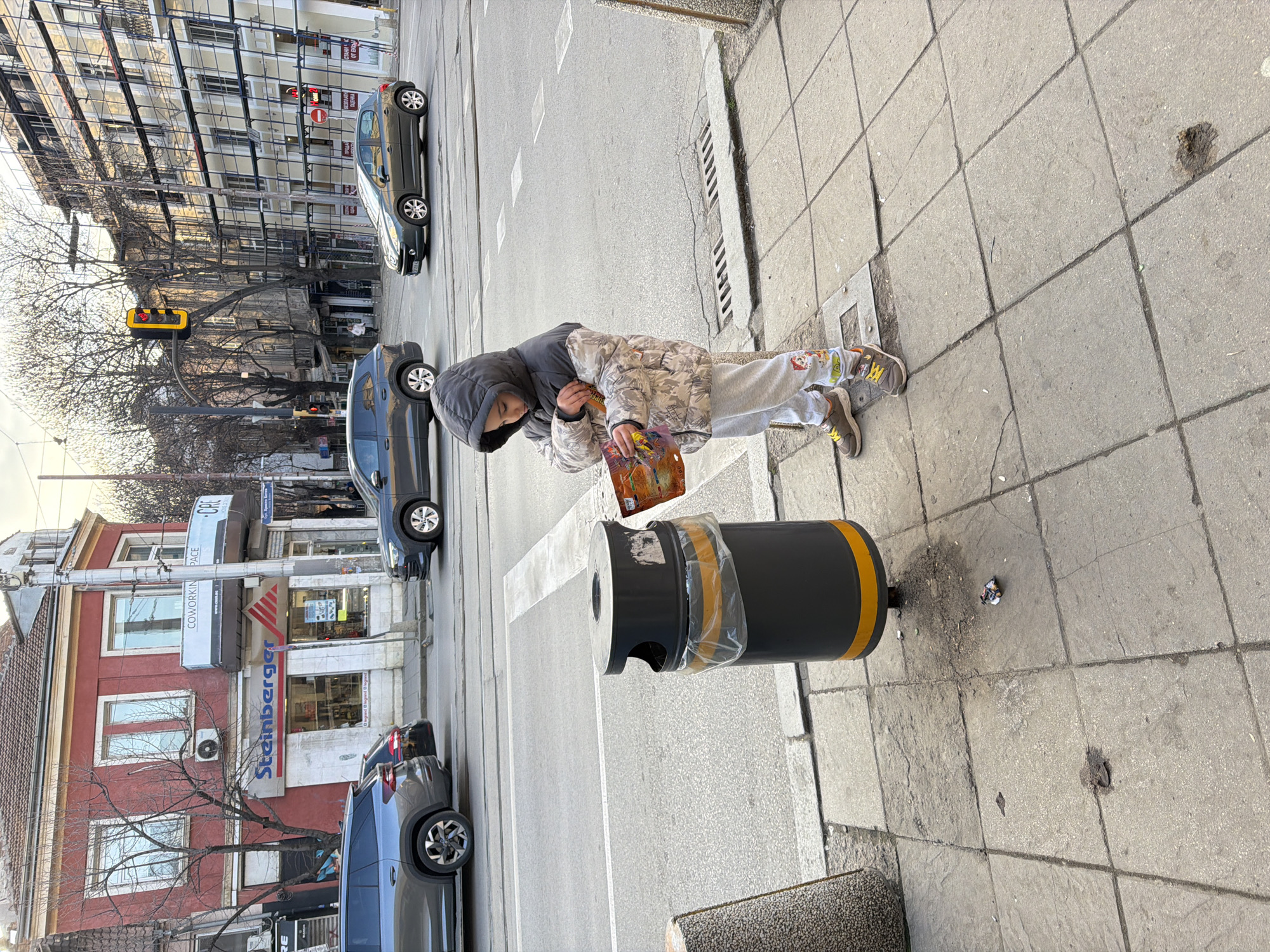 An outdoor urban scene in Sofia, Bulgaria, specifically on Bul. Hristo Botev, depicts a young child on a cracked gray tiled sidewalk next to a dark gray public trash bin. The child, wearing a dark gray hooded jacket over a camouflage-patterned quilted jacket, light gray sweatpants with "Paw Patrol" characters, and brown and yellow sneakers, intently examines a colorful packet held in their hands. Small pieces of litter are visible on the pavement near the trash bin.
Beyond the sidewalk, an asphalt street with white road markings and apparent tram lines accommodates several vehicles: a dark gray SUV in the foreground, another dark vehicle behind it, and a dark gray hatchback moving in the opposite direction further down the street. A metal drainage grate is visible in the road.
The background features multiple buildings. To the left, a two-story red building with a tiled roof houses a "Steinberger" store and a "COWORKING SPACE - CRE." To the right, a multi-story building is covered extensively in scaffolding, indicating construction or renovation. Bare deciduous trees are interspersed among the buildings, and a traffic light displaying a red signal is suspended over the street. The sky is overcast.