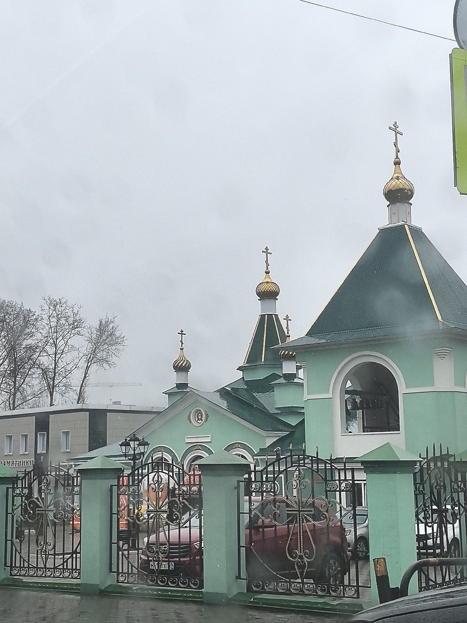 An outdoor, slightly wet-looking image captures a light green Russian Orthodox church under an overcast sky. The church features multiple golden onion domes, each topped with a distinctive Orthodox cross. The main part of the church is painted in a pale green color with darker green roofs, accented with golden trim. To the right, a bell tower-like structure with an arched opening is visible.
In the foreground, a decorative black wrought-iron fence, incorporating stylized crosses into its design, separates the viewer from the church grounds. The fence is set between light green pillars that match the church's color. Behind the fence, a red car is partially visible, and its license plate appears to show "M11 T 12" (likely a Russian plate, with '12' as a region code). Another dark vehicle is partially visible in the bottom right corner, suggesting the photo was taken from inside a car or from a street.
In the mid-ground, above an entrance or window on the church, there is a round icon or painting of a religious figure.
The background shows bare-branched trees, suggesting a colder season or early spring. Behind the trees and the church, a modern, light-colored building with several windows can be seen. Partially visible text on this building appears to be in Cyrillic, reading "...ПАМЯТНИКИ". A construction crane is also faintly visible in the distance, behind the trees.
The overall scene is subdued due to the gray, cloudy weather. There are no people visible, and no specific activity is taking place beyond the static presence of the church and surrounding environment. The image conveys a calm, somewhat solemn atmosphere typical of a cloudy day.