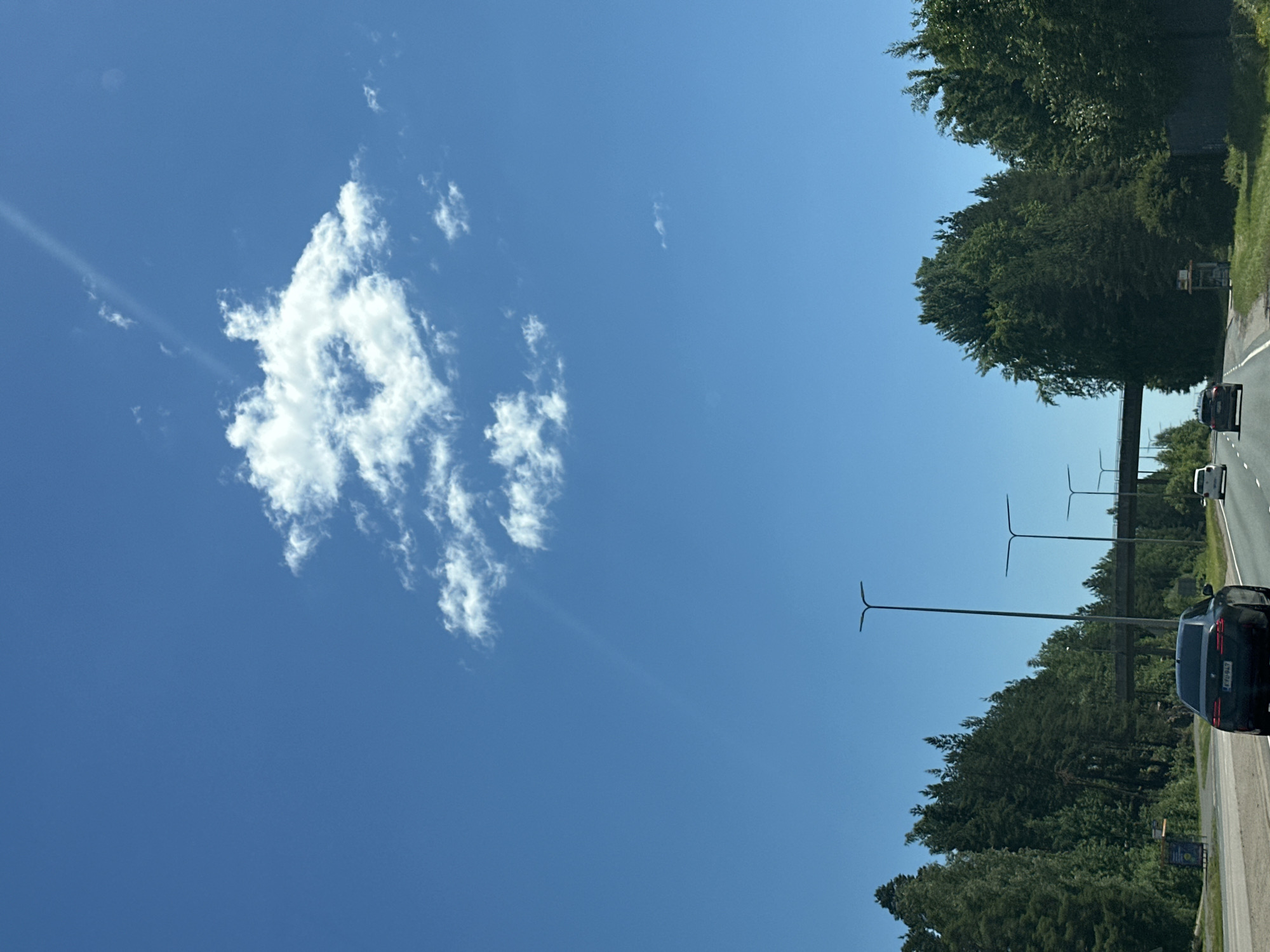 The image shows a view from a car driving on a highway on a clear day. The sky is a bright blue with a few scattered white clouds. A single, large cloud is prominent in the upper center of the frame, with a faint sunbeam passing through it. A thin, diagonal line, possibly a contrail or another sunbeam, streaks across the sky from the upper left to the lower right.
In the foreground, the asphalt highway is visible with lane markings. A dark-colored sedan is in the left lane, with its license plate partially visible. To the right of the sedan, a white SUV is further down the road. In the distance, there are several streetlights with their distinctive T-shaped fixtures. A bridge or overpass crosses the highway in the middle ground.
The sides of the highway are lined with dense green trees, indicating a possibly rural or suburban setting. On the left side, the trees are tall and coniferous, interspersed with some deciduous trees. On the right, the trees are also dense and green. A small, cubical structure with a transparent front panel and a dark roof sits near the road on the right side, possibly a bus stop shelter or information kiosk. The overall scene depicts travel through a tree-lined highway under a clear, sunny sky.