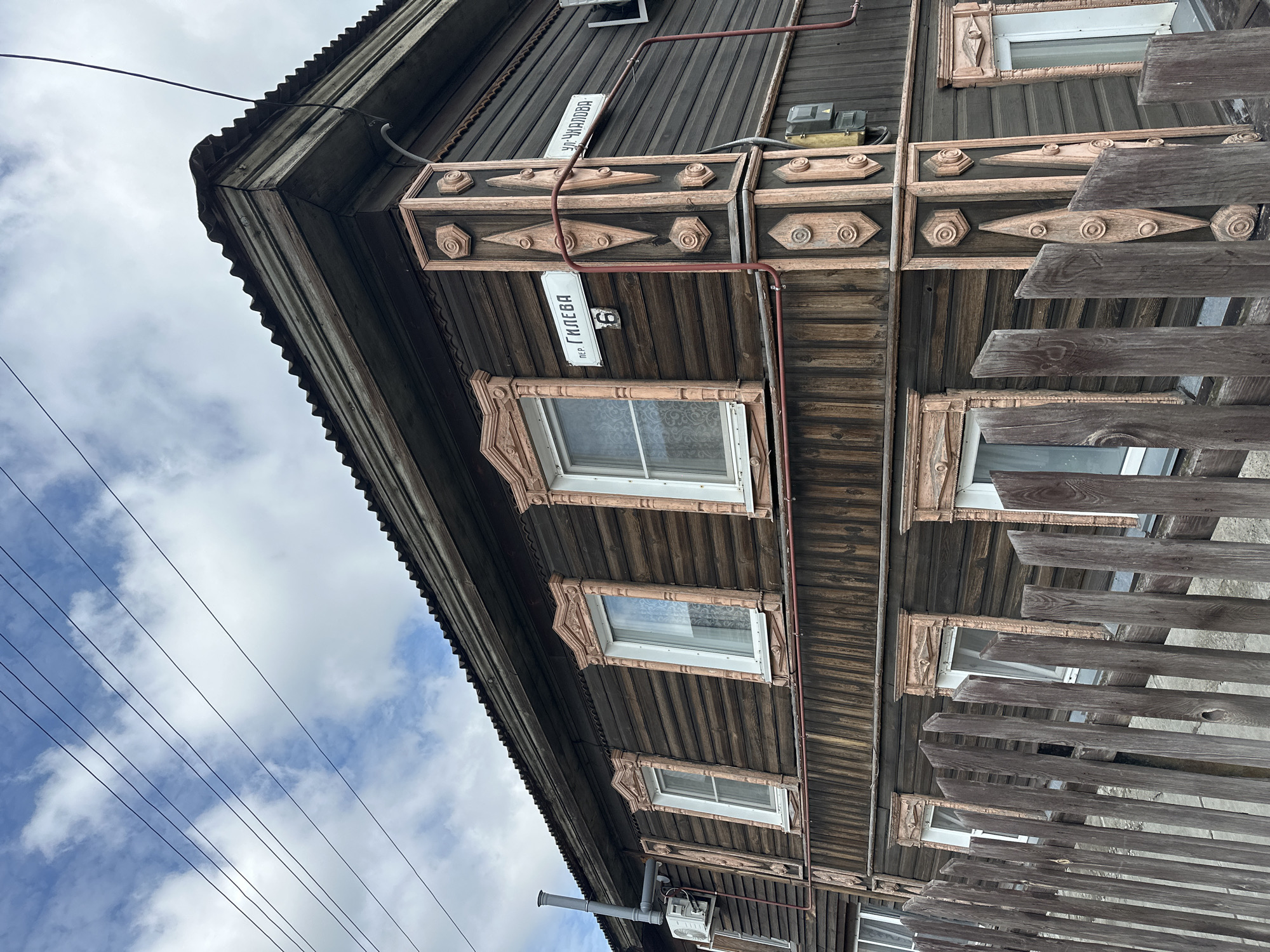 The image shows a close-up view of the corner of a two-story wooden house in the city of Nogornyi, Russia. The house is clad in dark horizontal wooden planks and features ornate pink-colored wooden trim around the windows and on the corner. Two street signs are visible on the corner post: one reads "пер. ГИЛЕВА 6" (Pereulok GILEVA 6, meaning GILEVA Lane, house number 6) and the other reads "ул. ЧКАЛОВА" (Ulitsa CHKALOVA, meaning CHKALOVA Street). A rustic wooden fence runs along the bottom of the image. The sky above is cloudy with visible power lines crossing the frame. An air conditioning unit and a ventilation pipe are mounted on the side of the house. The overall scene depicts a residential building in a Russian urban or suburban setting.
