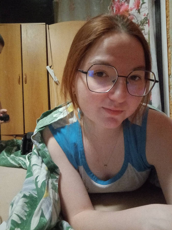 The image displays a young female, with light reddish-brown hair and black octagonal-framed eyeglasses, lying prone and propped on her forearms. She is looking directly at the camera with a slight smile. Her attire consists of a blue and white sleeveless top, and a thin gold-colored necklace with a small pendant is visible around her neck. A green blanket adorned with a white floral and leaf pattern covers her lower body and left arm. In the blurred background, to the upper left, the crown of another individual's head and their right shoulder are partially visible, holding a dark object. Behind the subjects, two light brown wooden wardrobe units with dark handles are present, alongside a white charging adapter or similar device with a visible cable hanging on the wall between them. A dark electronic device, possibly a camera or phone, is situated on the surface to the lower left, partially obscured by the blanket. The scene is an indoor residential setting in the city of Старый Шагирт, Russia.
