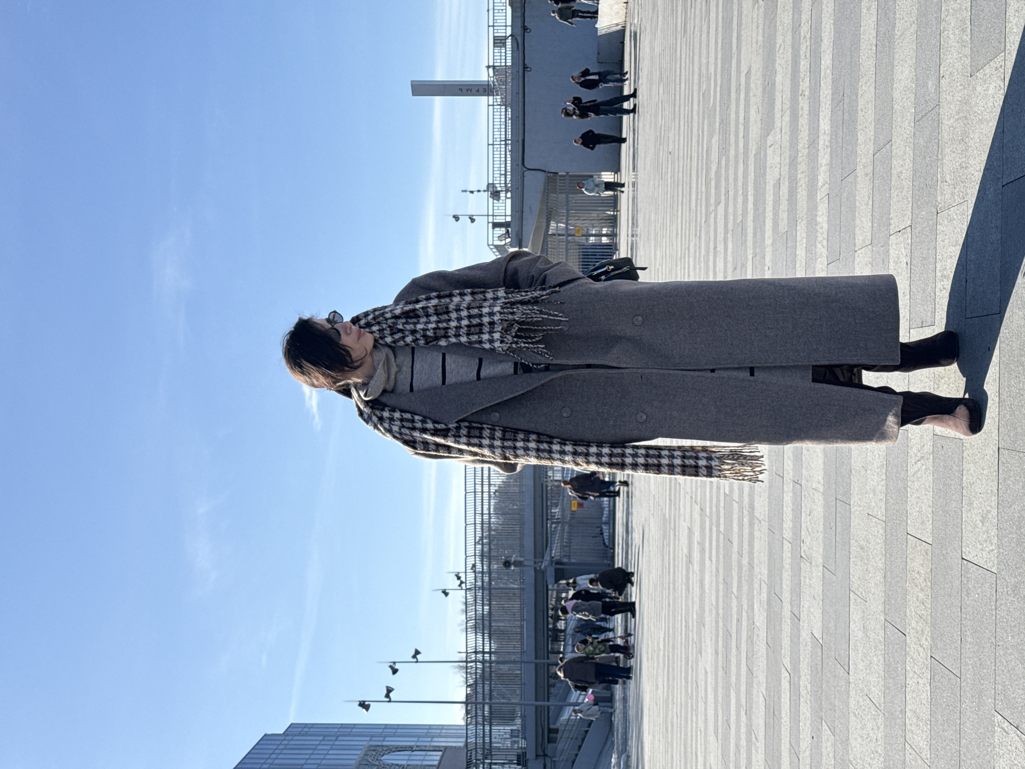 A woman stands in a paved public square under a clear blue sky in Perm, Russia. The individual, centrally positioned, has dark hair, wears dark sunglasses, a light grey striped turtleneck, a long grey overcoat with buttons, a dark and light patterned scarf draped around her neck, and dark boots. Her hands are in her coat pockets, and her head is turned to her right (viewer's left). A distinct shadow from her body is cast diagonally on the ground in front of her.
The ground is composed of light and dark grey rectangular paving stones arranged in a striped pattern. In the mid-ground, a pedestrian overpass with metal railings crosses horizontally. To the left, a modern building with a glass facade is visible. Numerous pedestrians are present in the background, walking or standing on various levels of the square and the overpass. One individual in the far-right mid-ground appears to be holding a camera or phone. A child on a scooter is visible among other people on the left side of the mid-ground. Streetlights with multiple lamp heads are visible on poles.
On the far right of the background, a tall, light-colored rectangular monument column displays the vertical Cyrillic inscription "ПЕРМЬ" (Perm), identifying the location as the Perm-1 railway station square, near the Perm River Station. The scene is brightly lit by direct sunlight.