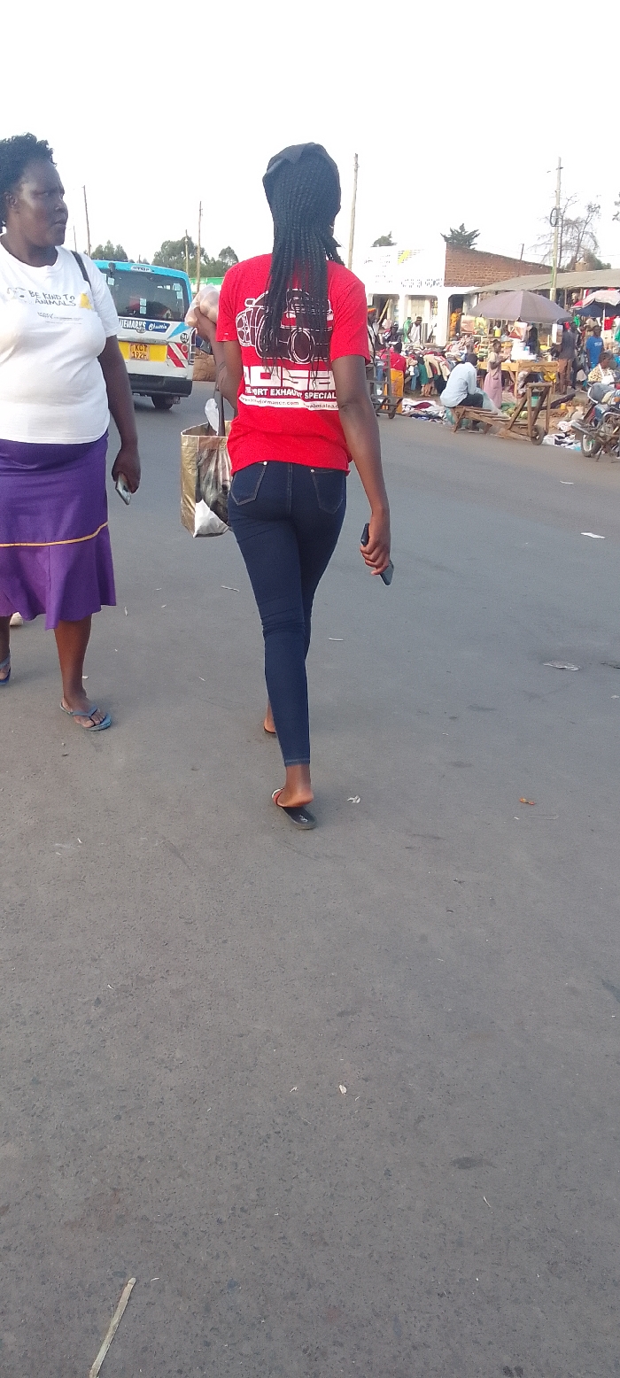 The image depicts an outdoor street scene in Kakunga, Kenya, bustling with activity, particularly in the background which suggests a market environment. The time of day appears to be bright, possibly morning or late afternoon, with a clear sky overhead.
Two women are the primary subjects in the foreground, walking along a gray asphalt road. The woman closer to the camera is seen from behind, walking away. She has long, dark braided hair and is wearing a red short-sleeved t-shirt, dark blue jeans, and black flip-flops. Her t-shirt displays a distinctive graphic of a camera, seemingly integrated with an automotive exhaust pipe design. Text on her shirt includes "OGE", "EXHAUST SPECIAL", "www.performance.com", and "salsaa.com". She carries a shiny shopping bag in her left hand and a dark-colored object, likely a phone, in her right.
To her left, another woman walks partially facing the camera. She has short, dark curly hair and is dressed in a white short-sleeved t-shirt, a purple skirt with a yellow or orange trim, and blue flip-flops. Her t-shirt bears the text "BE KIND TO ANIMALS". She is also holding a light-colored object, possibly a phone, in her right hand.
In the mid-ground, a light blue and yellow public transport vehicle, commonly known as a matatu, is visible. The words "NYEMARK'S Classic" and a Kenyan license plate "KCQ 122H" can be identified on its side. The background on the right reveals a vibrant market area with numerous people, stalls, and goods displayed on the ground, under makeshift shelters and simple buildings. The overall mood is one of everyday life and commerce. No specific emotions are evident from the subjects.