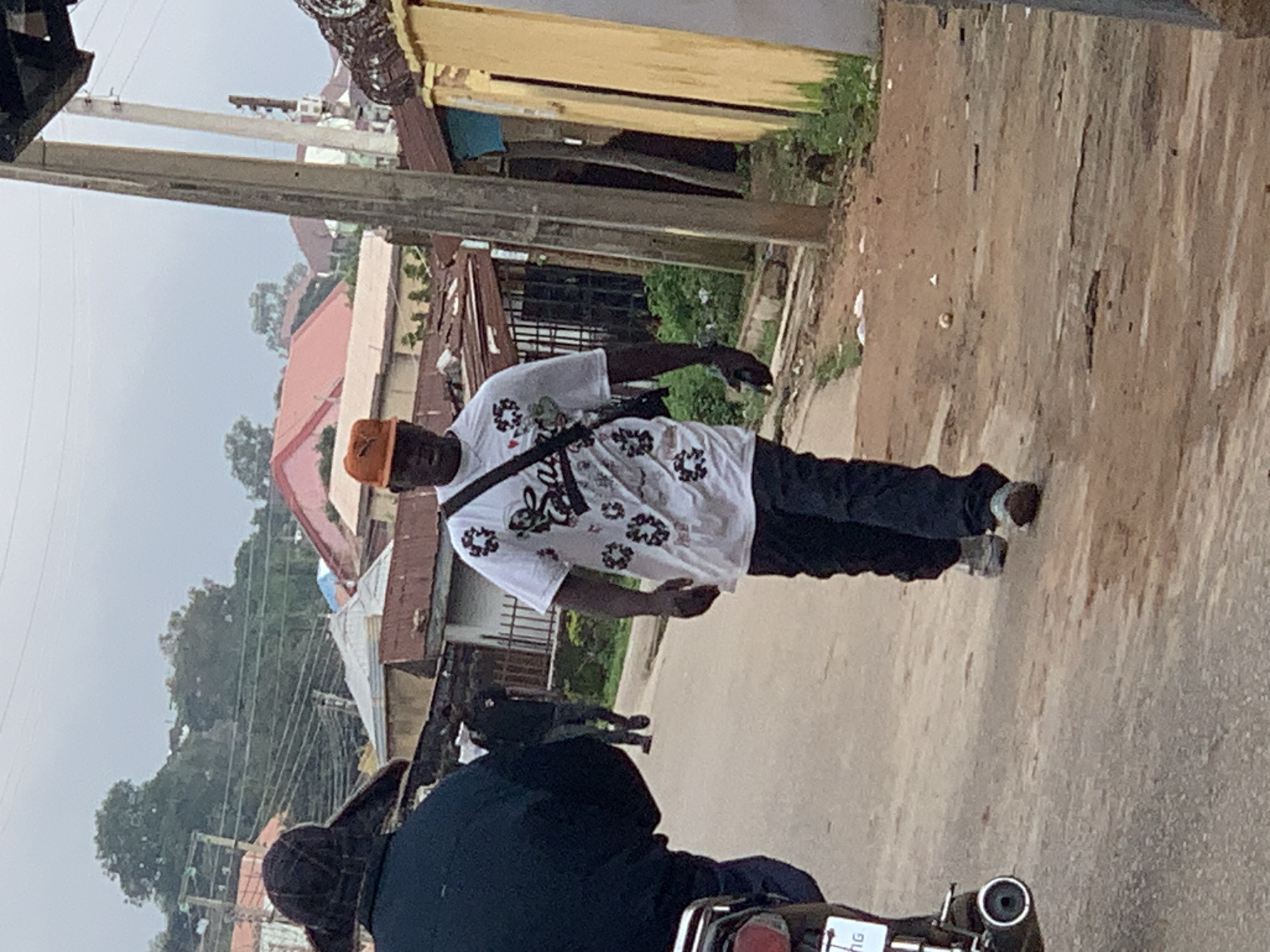 A medium shot captures a street scene in Jos, Nigeria, with a man in the foreground walking towards the camera. He wears a white t-shirt with a graphic print, dark pants, and an orange baseball cap. A black strap crosses his chest, holding a dark bag. His right hand is extended, possibly holding a mobile phone. To his left, another person, partially visible from behind, is wearing a dark jacket and a patterned hat. In the background, a second man walks away from the camera down the street, dressed in dark clothing. The environment features a dusty road, modest buildings with corrugated iron roofs, electrical poles, and overgrown vegetation. The sky is overcast, suggesting a cloudy day.