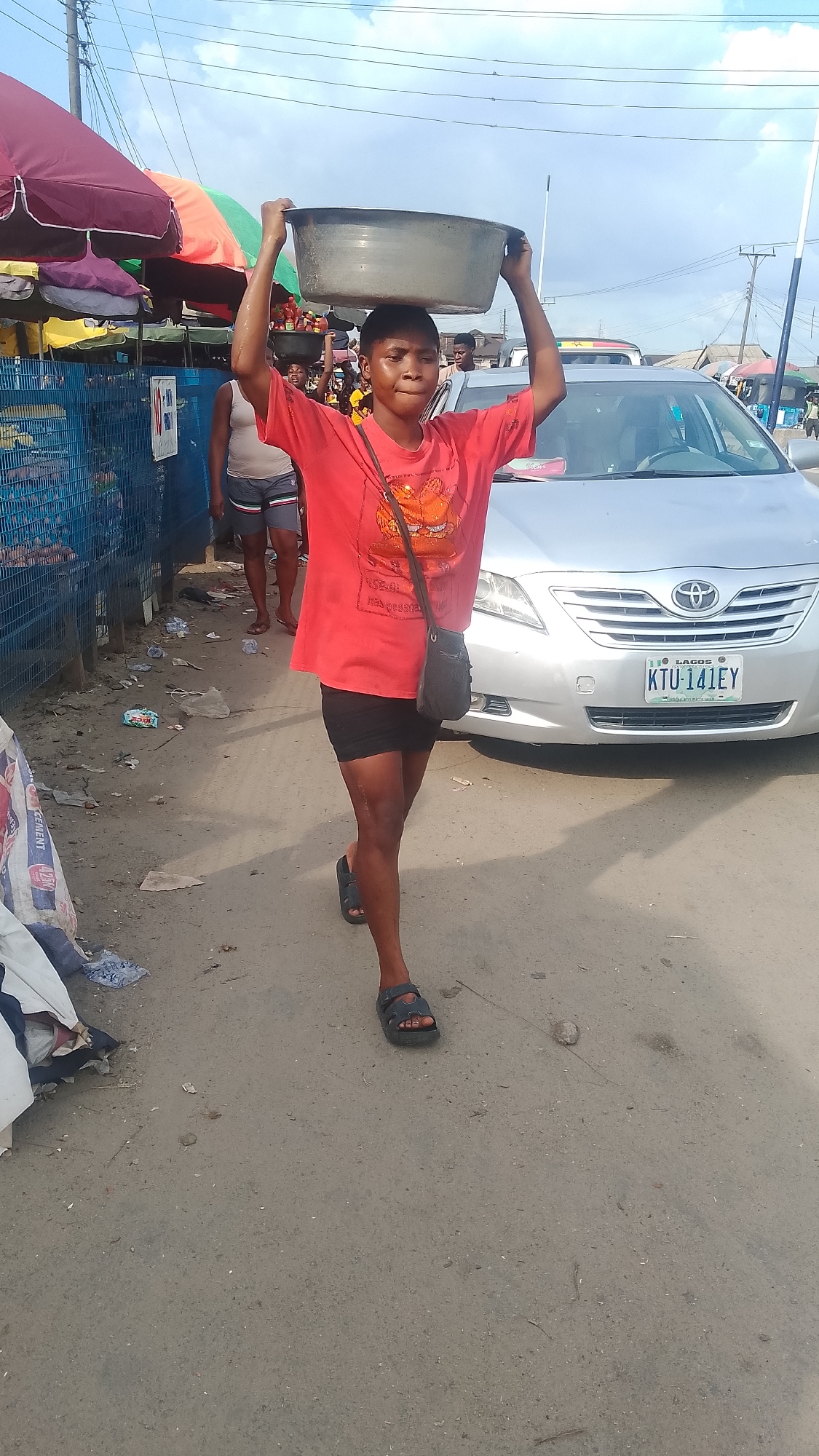 A person in a red t-shirt and black shorts carries a large gray bowl on their head while walking on a dirt and paved road in Warri, Nigeria. The person is wearing sandals and has a black bag slung over their shoulder. A silver Toyota car with the license plate "KTU-141EY" is parked behind the person. There is trash scattered on the side of the road. People are visible in the background, along with market stalls under colorful umbrellas. Overhead power lines are visible in the sky. The picture seems to be taken during daylight.