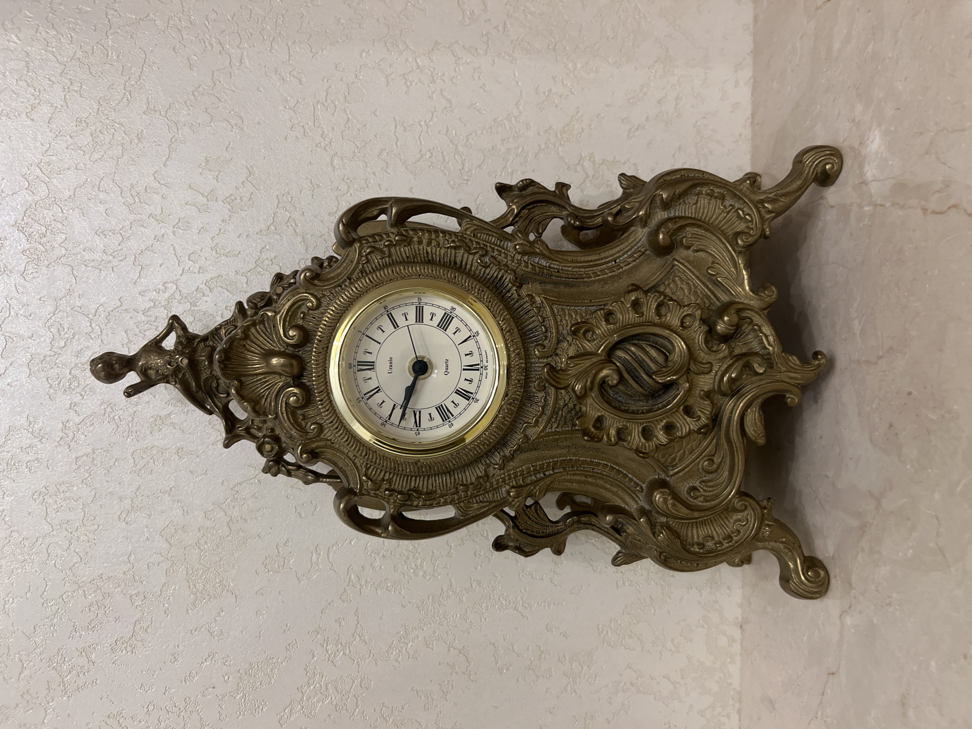 A detailed, close-up, front-facing shot showcases an ornate, antique brass mantel clock with a white face and Roman numerals. The clock features elaborate scrolling, filigree, and a small figural element at the apex. It is positioned on a marble-like surface, with a textured, off-white wall as the background. The clock's hands indicate a time of approximately 2:23. There are no people present in the image, and the environment appears to be indoors, likely a domestic setting in Bender, Moldova. The lighting is diffused, highlighting the metallic sheen of the clock and the subtle texture of the wall.