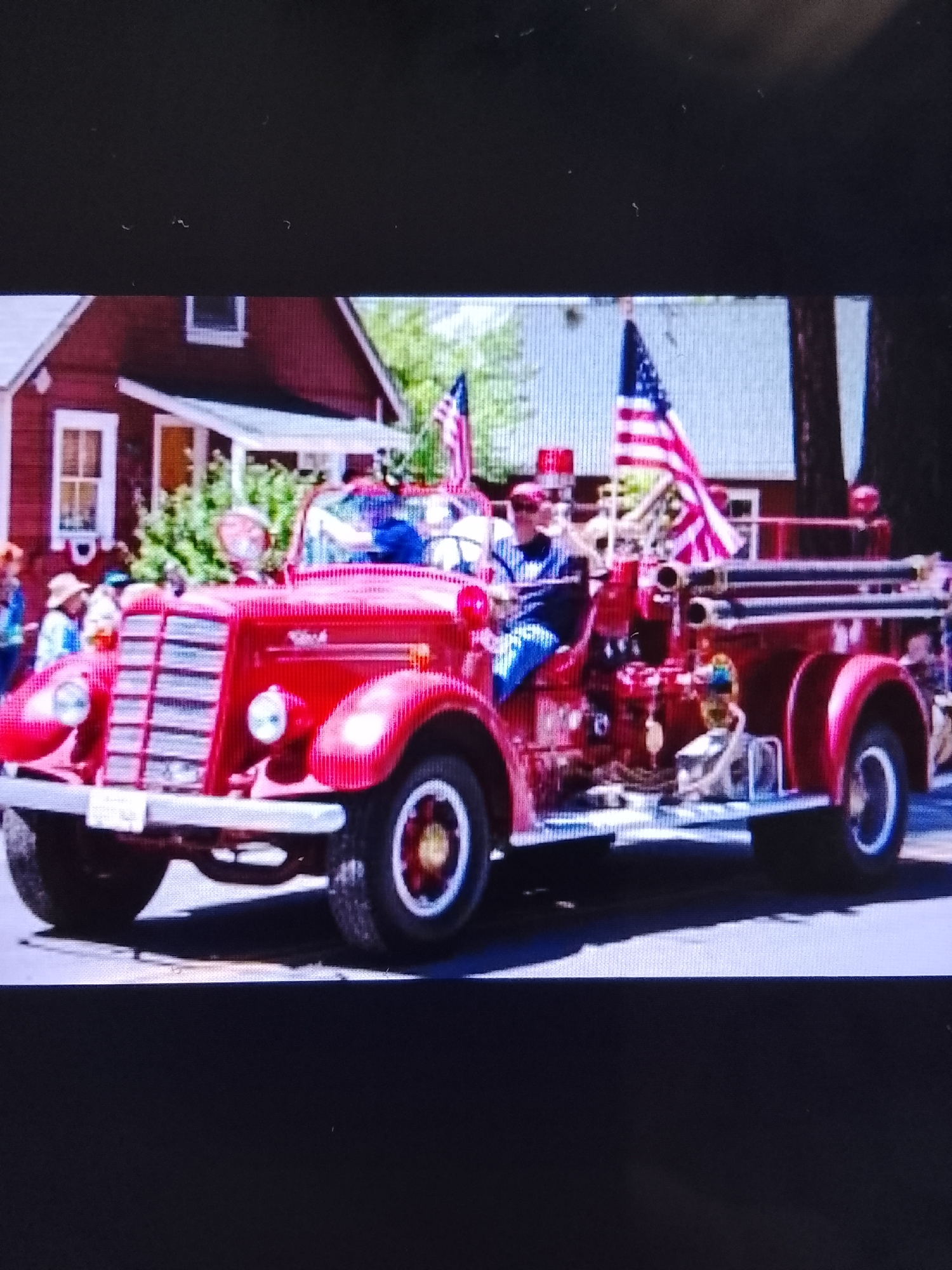 This is a photograph of a vintage red fire truck participating in a parade. The fire truck is adorned with American flags and has people riding in it. In the background, a red building with white trim is visible, along with some greenery. The scene appears to be outdoors, likely on a street or in a town setting. The question asks for the location in Safonovo, Russia, but there is no information in the image or the provided crops that would indicate this specific location.