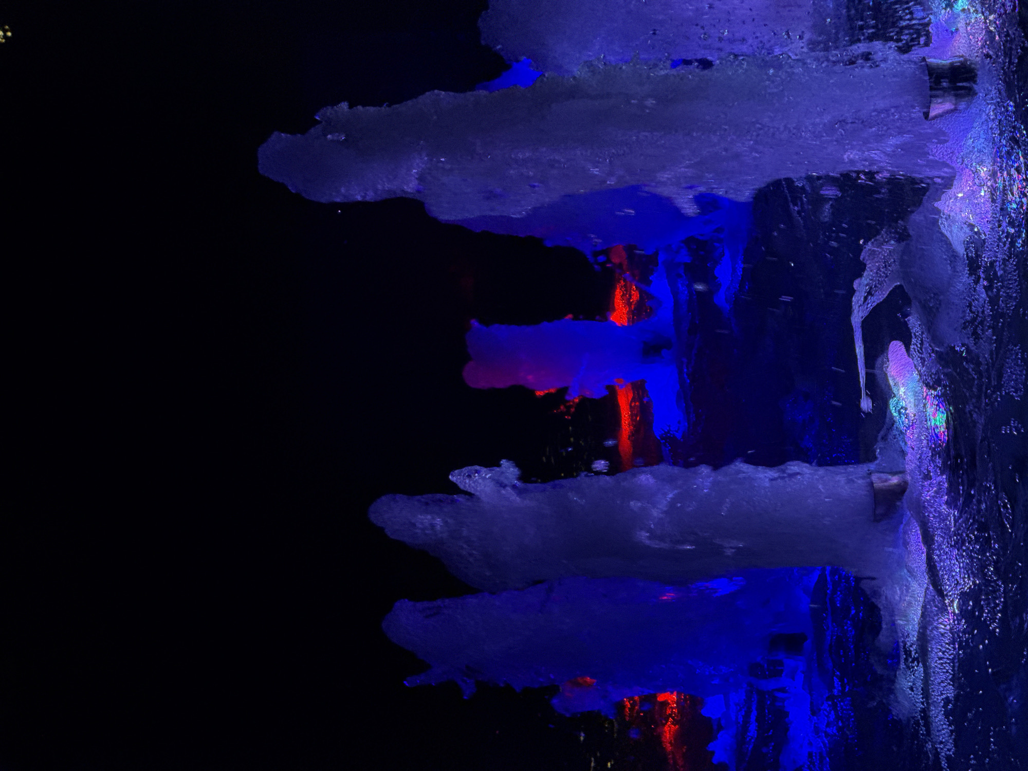 A nighttime photograph captures several illuminated water fountains in the city of Unknown, Unknown. The water jets are lit with vibrant blue and some red light, creating a surreal and dramatic effect against the dark background. The water appears to be in motion, with some streams frozen mid-air and others splashing into the pool below. The overall scene is abstract and artistic, focusing on the interplay of light and water.