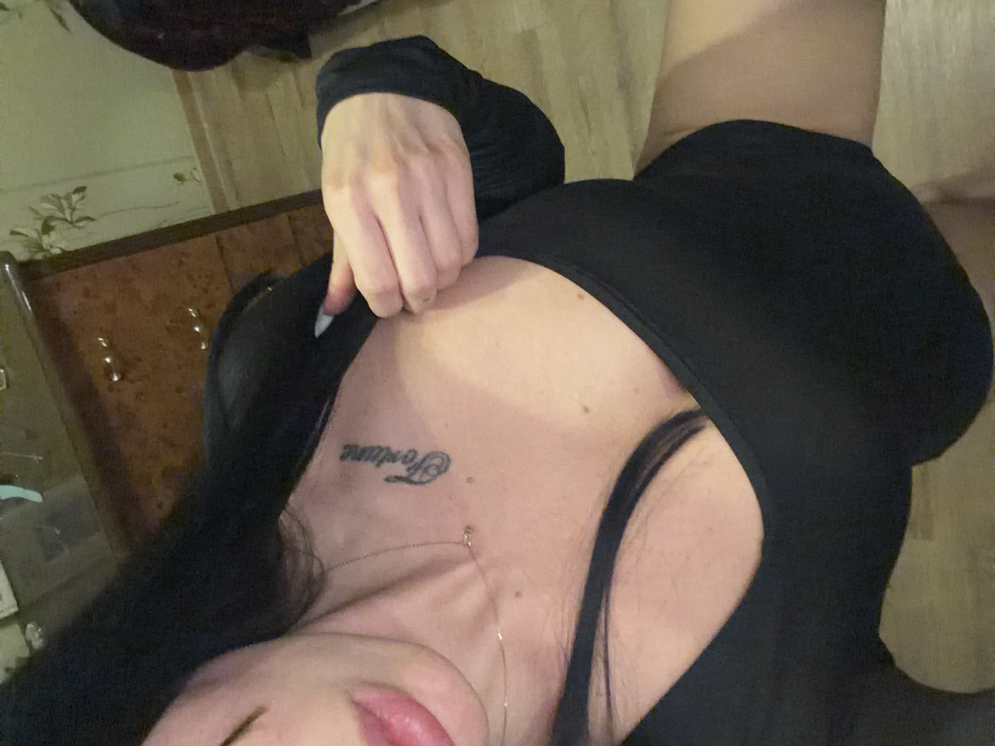 A person is lying down indoors, their upper body visible. They are wearing a black, long-sleeved top. A tattoo of the word "Fortune" is visible on their neck. The background shows wooden furniture and wallpaper.