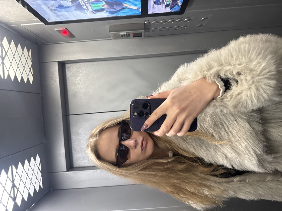 A young adult female is depicted taking a mirror selfie inside an elevator, located in the city of Unknown, Unknown. She has long, wavy blonde hair and wears dark, rectangular sunglasses. Her lips are slightly pursed. She is dressed in a light-colored, possibly off-white, faux fur coat over a dark garment. Her left hand holds a dark-colored smartphone, equipped with a prominent camera cluster, to capture the image. Her fingernails are painted a muted reddish-brown. The elevator interior features gray walls, a ceiling with a grid of recessed square lights, and a control panel on the right wall. This panel includes multiple buttons, a red indicator light, and a digital display screen exhibiting reversed text and an image of a person wearing ski goggles.