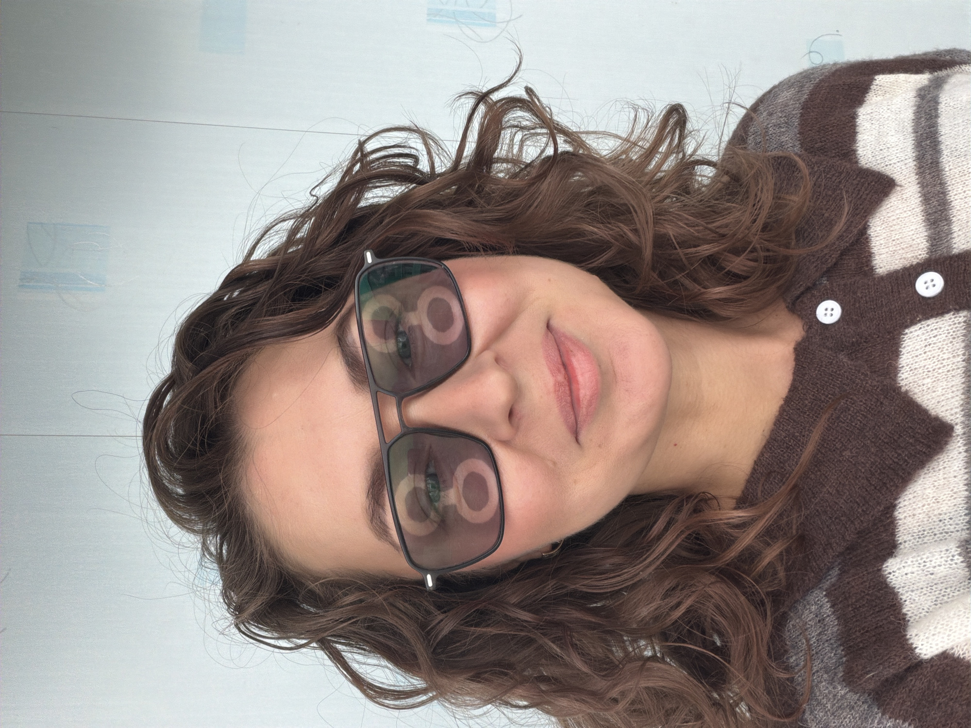 A woman with wavy brown hair and light skin is wearing sunglasses and a brown and white striped sweater with white buttons. The background appears to be a light-colored wall or floor. The location is Unknown, Unknown.