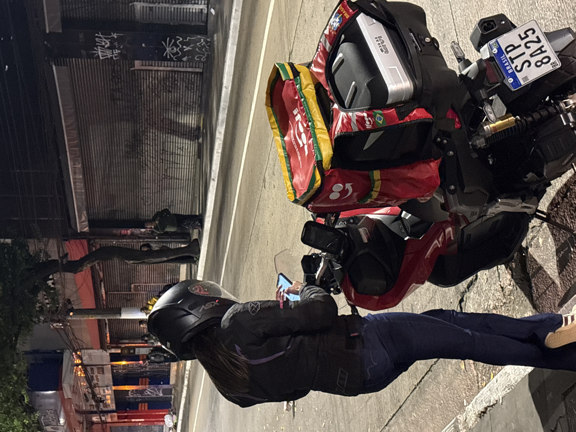 A person wearing a black motorcycle helmet and jacket stands on a sidewalk next to a red and black motorcycle. The person is looking at a smartphone in their hands. The motorcycle has a large red delivery bag on the back, with "Ifood" branding visible. A license plate reads "BRASIL STP 8A25 BR". In the background, a street with traffic lights and closed storefronts is visible. Graffiti adorns some of the buildings. The scene appears to be at night on a street in São Paulo, Brazil.