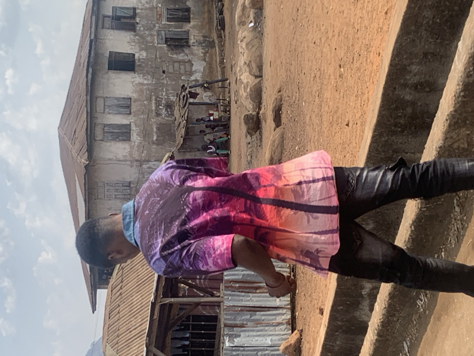The image depicts an outdoor scene in Egbedi, Nigeria, under an overcast sky. In the foreground, a male individual is walking away from the camera, positioned on a low concrete structure. He wears a short-sleeved collared shirt with a denim blue collar, featuring a gradient design from purple to pink to orange, adorned with dark palm tree silhouettes. He is also wearing dark, possibly distressed, jeans.
The ground throughout the scene is reddish-brown dirt, scattered with various rocks. To the left midground, a structure with a corrugated metal roof and walls is partially visible, with what appears to be laundry hanging inside. In the center midground, a makeshift shelter with a corrugated metal roof is present. Underneath this shelter, approximately five individuals, primarily men, are observed; some are seated, others standing, near a small white table or counter. A white standing fan is positioned outside the shelter.
In the background, a two-story concrete building with a visibly weathered and dirty facade and a rusted corrugated metal roof dominates the view. The building features multiple rectangular windows; some have closed wooden shutters, while others are open or appear empty. A distant hill or mountain is visible on the far left background.
