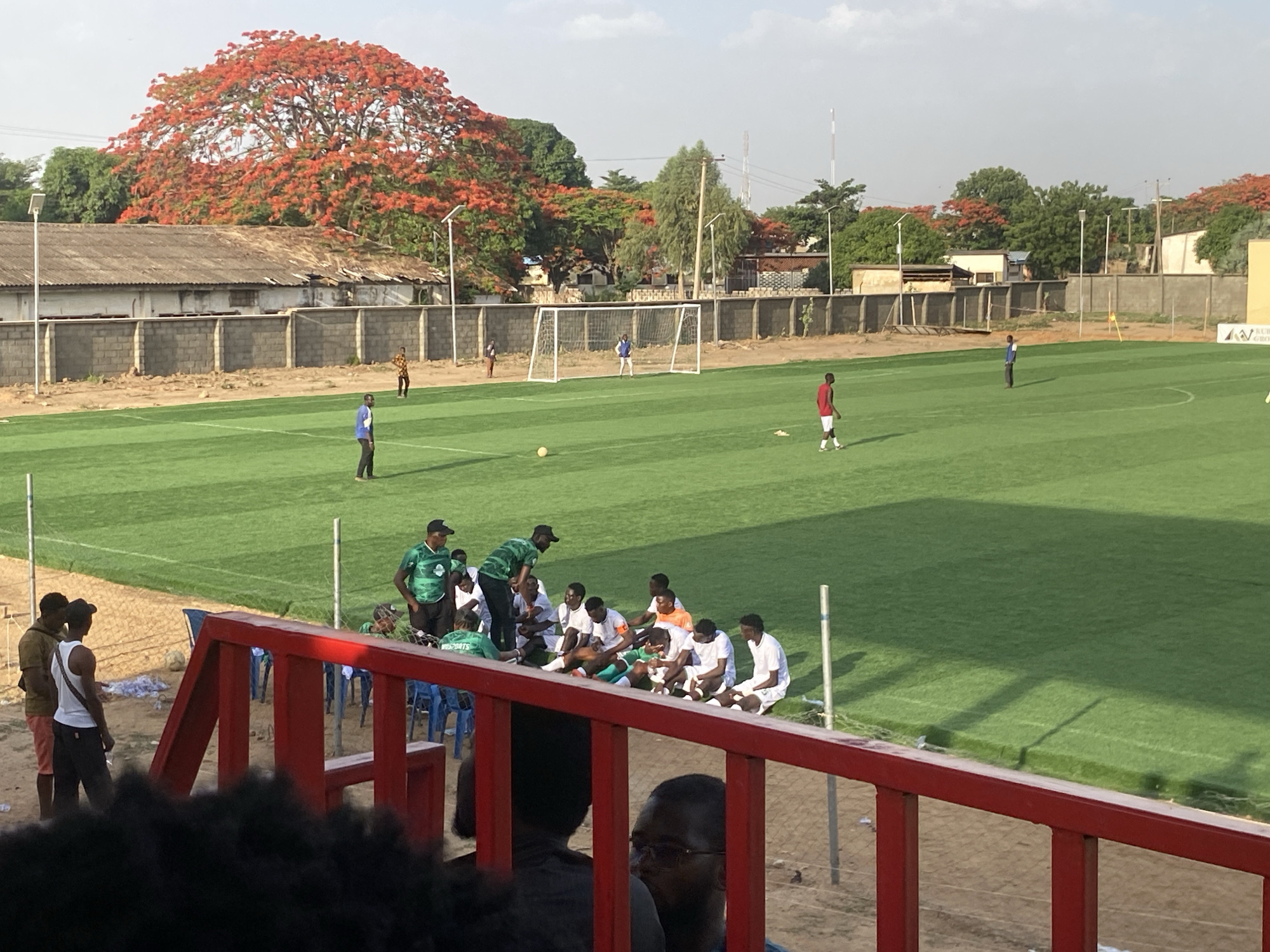 The image depicts a soccer field in Tudun Wada, Nigeria. The field is green and well-maintained, surrounded by a low wall and a mix of buildings and trees. A large tree with red flowers dominates the background. On the field, several people are engaged in soccer-related activities. A soccer goal is visible. To the right of the field, a group of individuals are seated on a bench. Two men are standing near the spectators. In the foreground, a red railing separates the viewer from the field. Some people are sitting behind the rail.