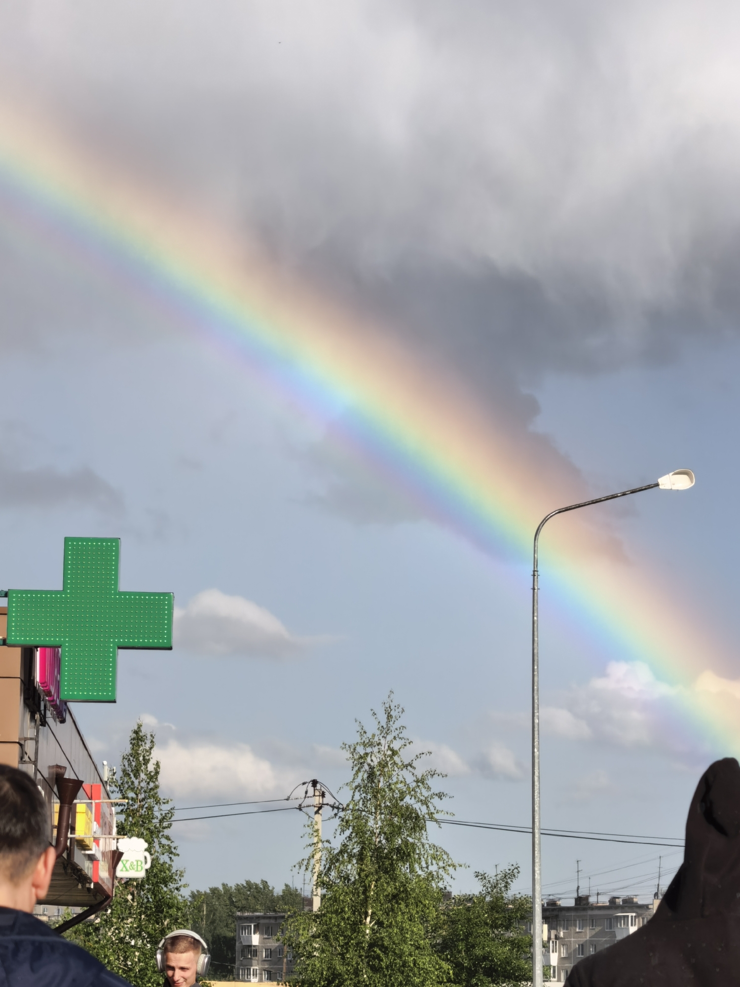 The image depicts a rainbow stretching across a cloudy sky, possibly in an urban area. In the foreground, there are several people visible, one wearing headphones. There's a green cross sign, possibly indicating a pharmacy or healthcare facility. Other details include a street light, trees, and buildings in the background. Electrical wires are visible. The overall impression is a somewhat overcast day with a notable rainbow appearing in the sky.