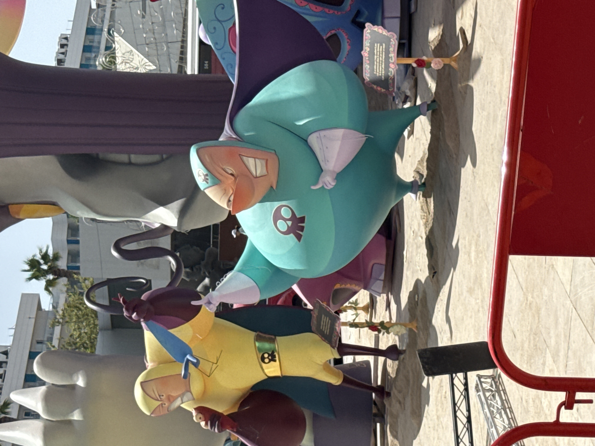 This is a medium-shot photograph of two cartoonish, larger-than-life figures dressed as superheroes. The figure on the left is wearing a yellow suit with a black belt and cape, and is holding a dark red, sack-like object in its left hand. It has an angry expression. The figure on the right is larger, wearing a teal suit with a purple cape, and also appears to have an angry expression. Both figures have skull symbols on their chests and masks. They are standing on sand, with a backdrop of modern buildings and palm trees. There are also various structures and what appear to be stage props or art installations around the figures. In the foreground, there is a red barrier. The scene appears to be outdoors, likely in a public space.